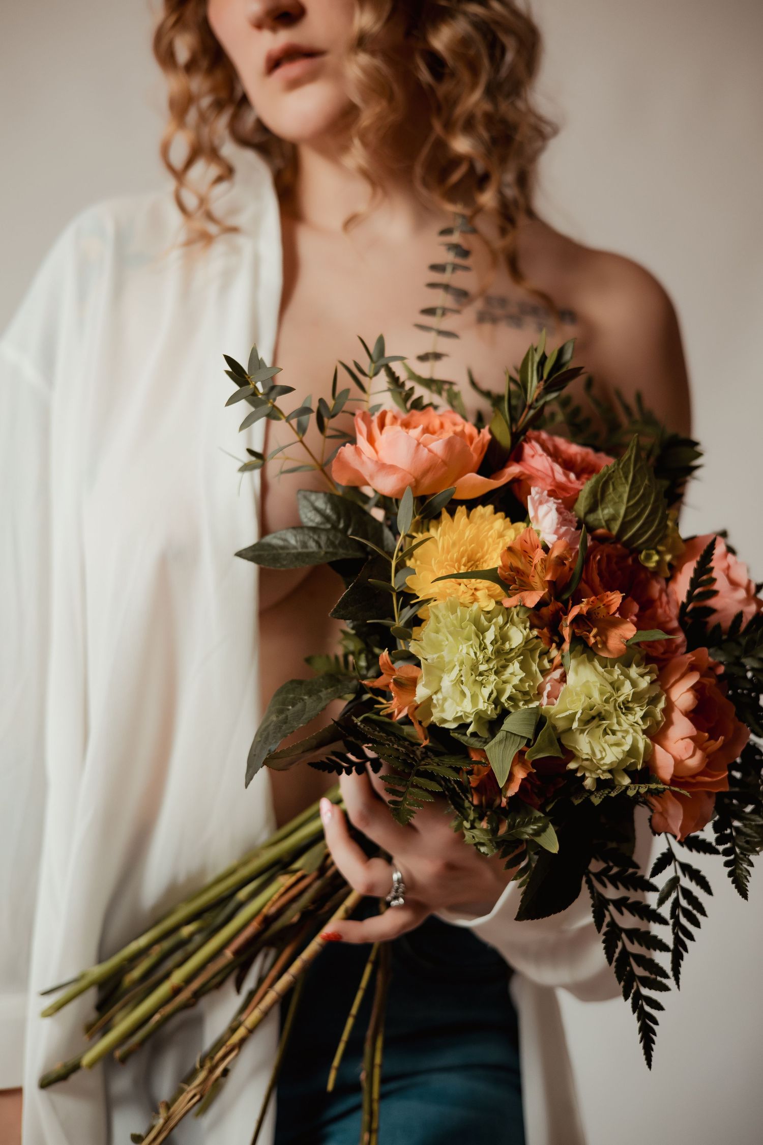 A person in a white top holds a vibrant bouquet of peach, yellow and green flowers with dark foliage against a neutral background.