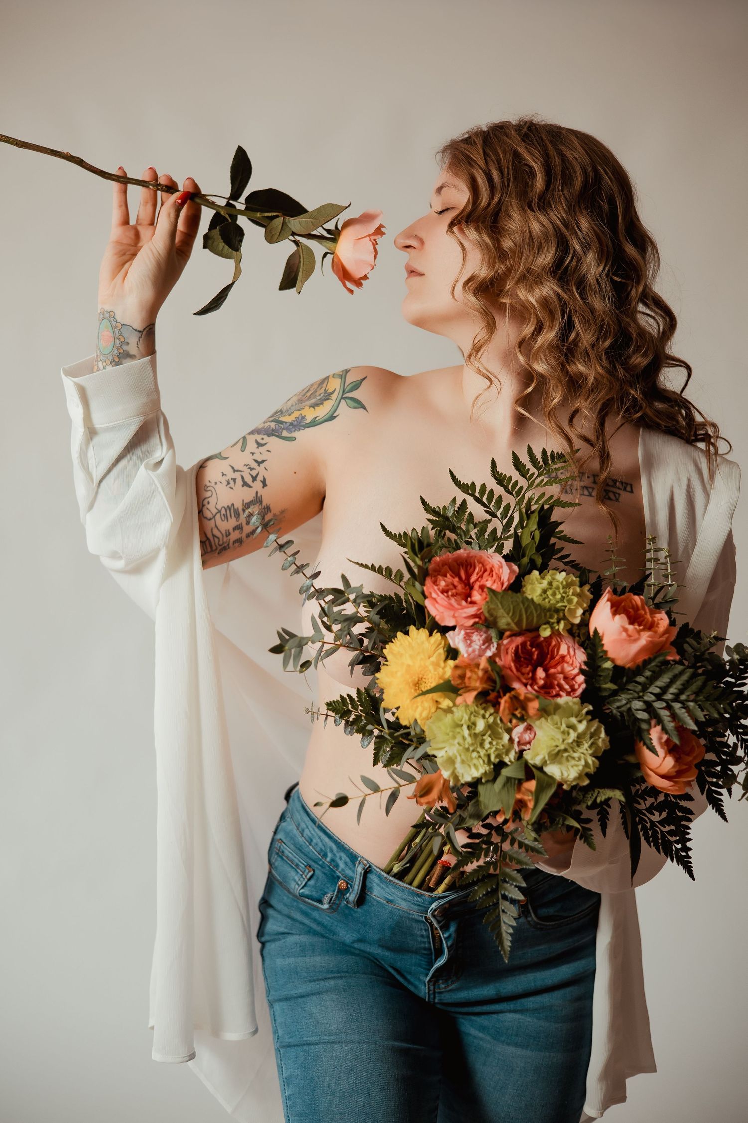 Artistic boudoir portrait featuring curly hair, tattoos, white shirt, jeans and colorful floral bouquet against neutral wall.