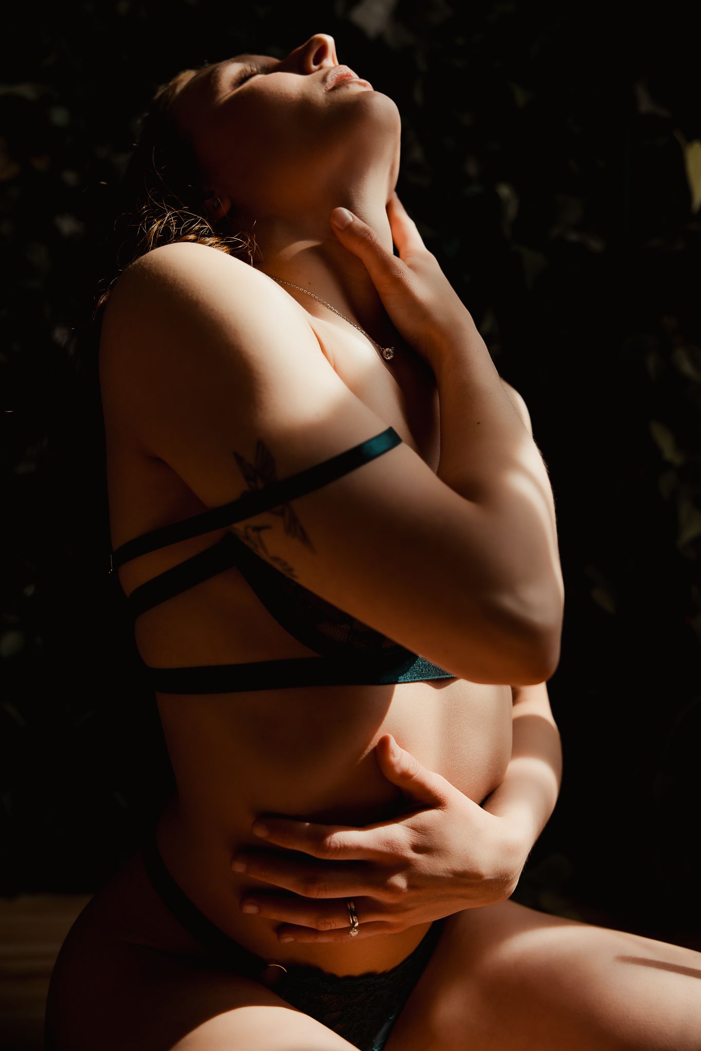 Artistic boudoir photo with dramatic shadows showing crossed black straps against bare skin in natural lighting.