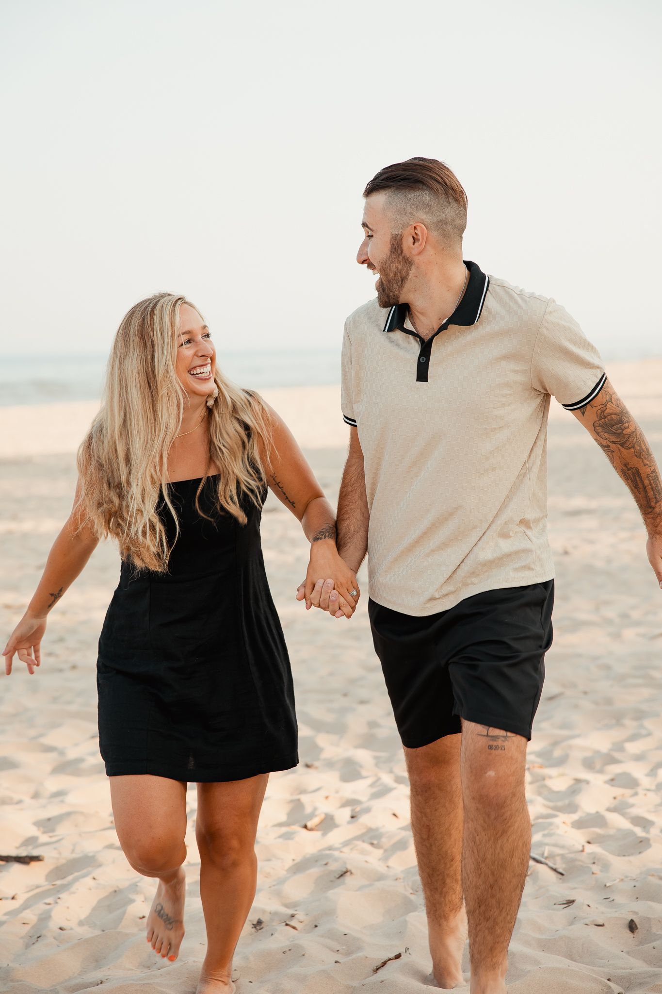 A couple walking playfully together on a sandy beach wearing matching black and beige outfits at sunset.
