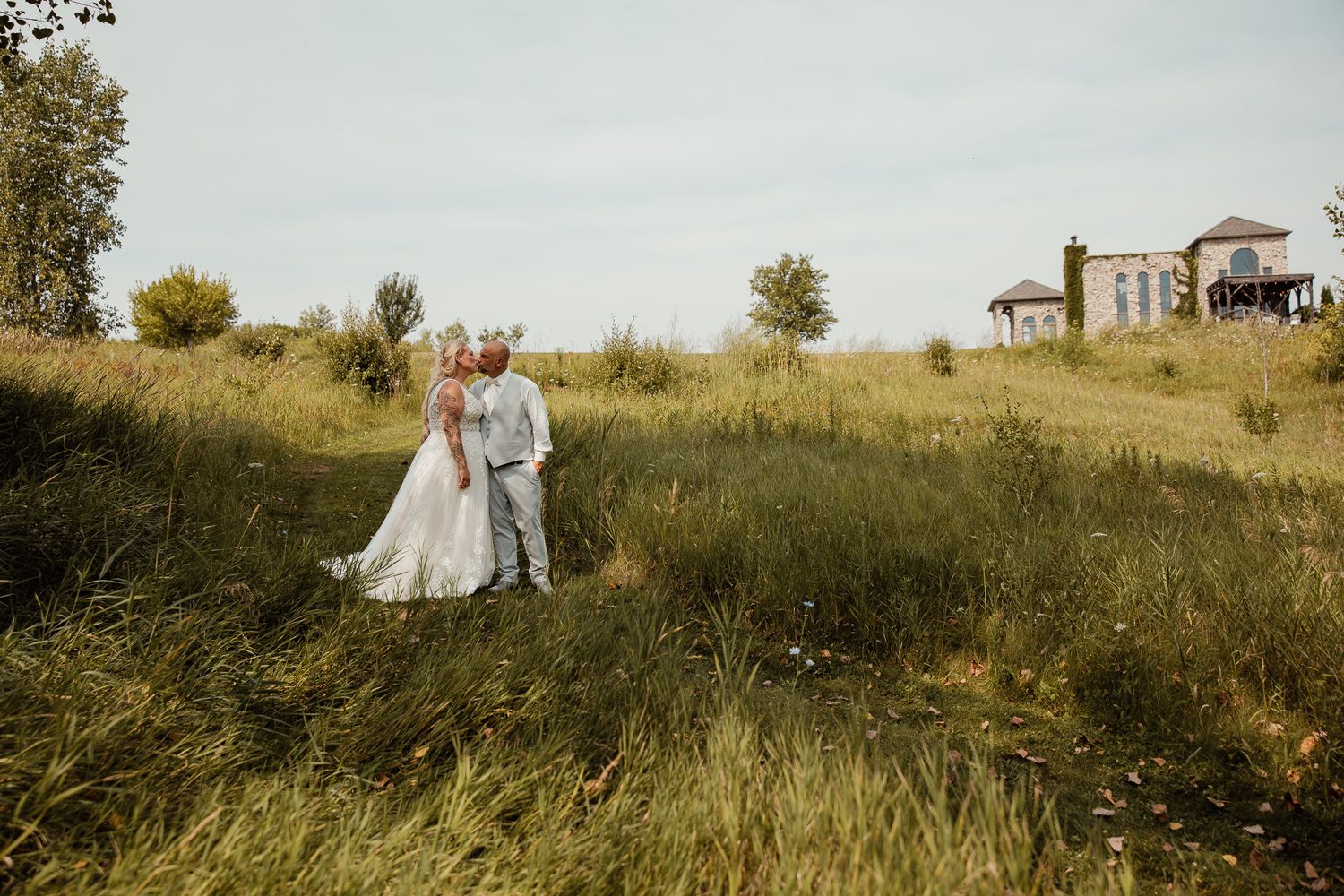 Romantic outdoor wedding photos taken in a golden field with a rustic building in the background at sunset.