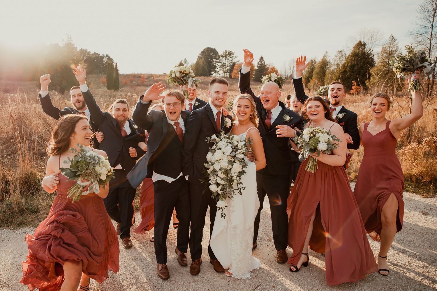 Wedding party celebrates outdoors with raised hands wearing burgundy attire at sunset. wedding