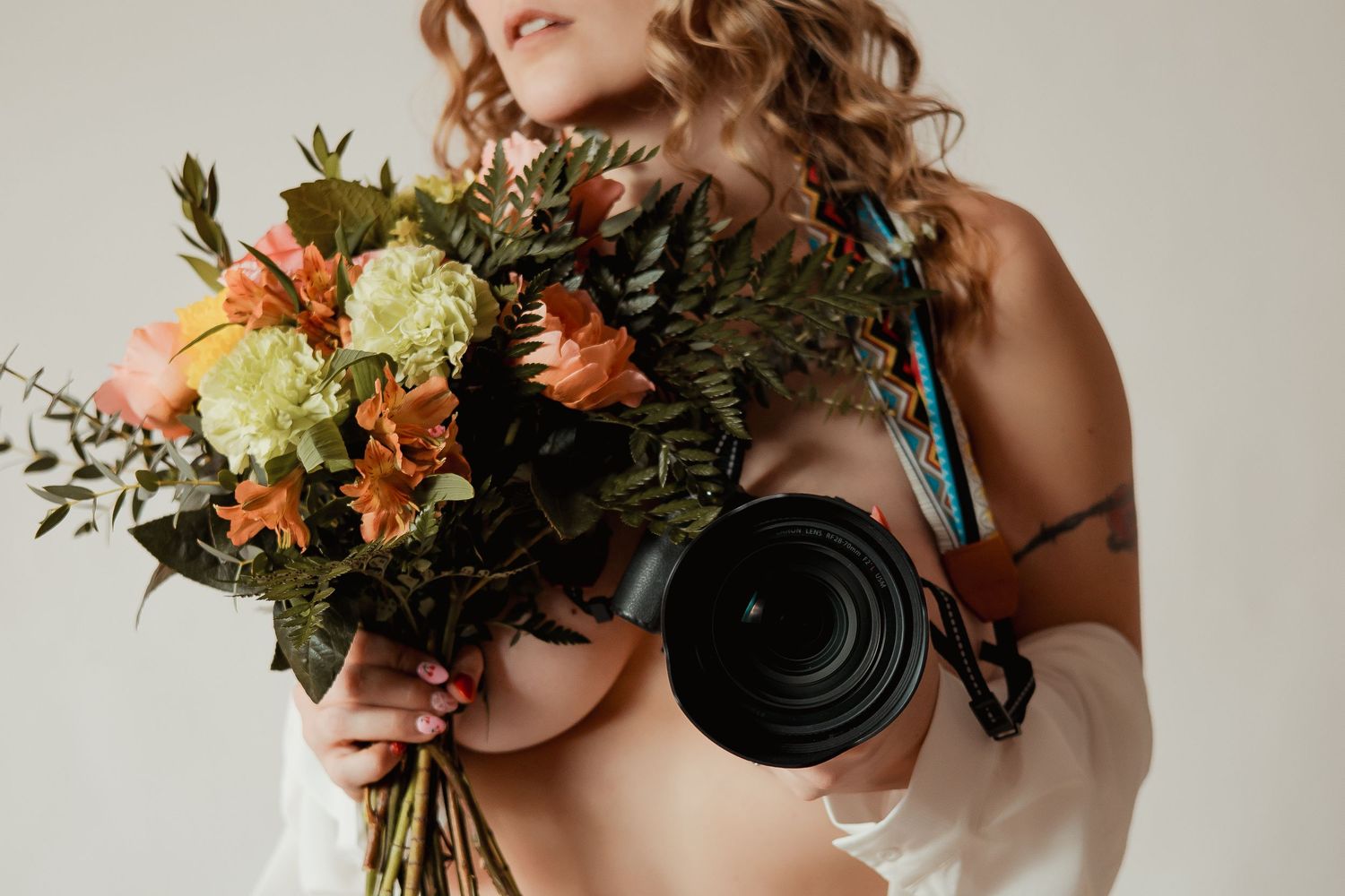Photographer holding professional camera and vibrant bouquet with orange and green flowers against white wall.