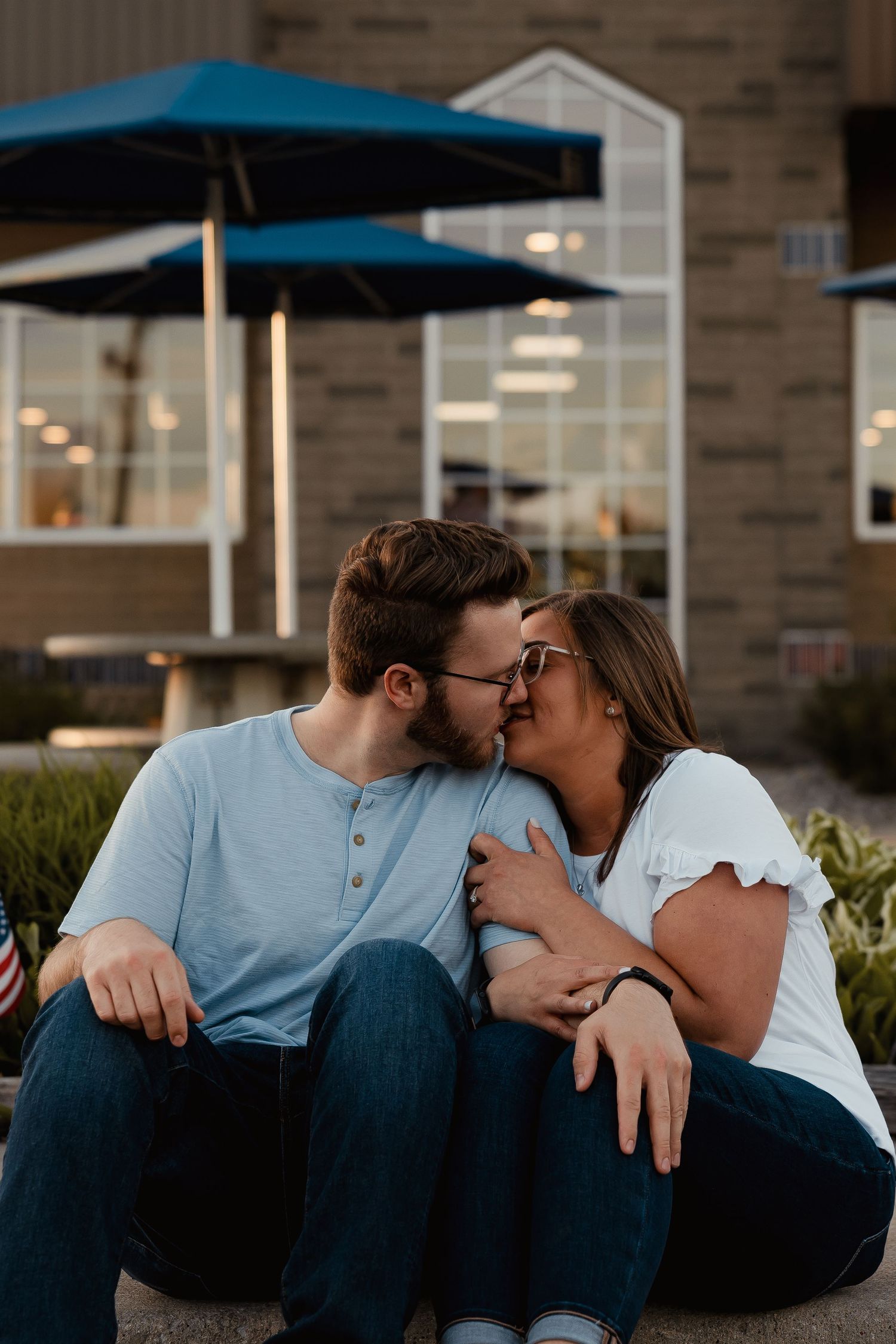 A couple shares a romantic kiss while sitting outdoors near a building with blue patio umbrellas at sunset.