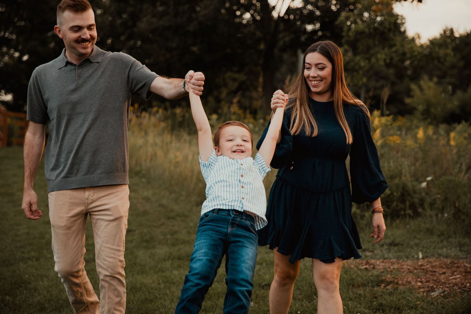 A happy family of three walks outdoors, parents swinging their young son by the hands in a lush green park.