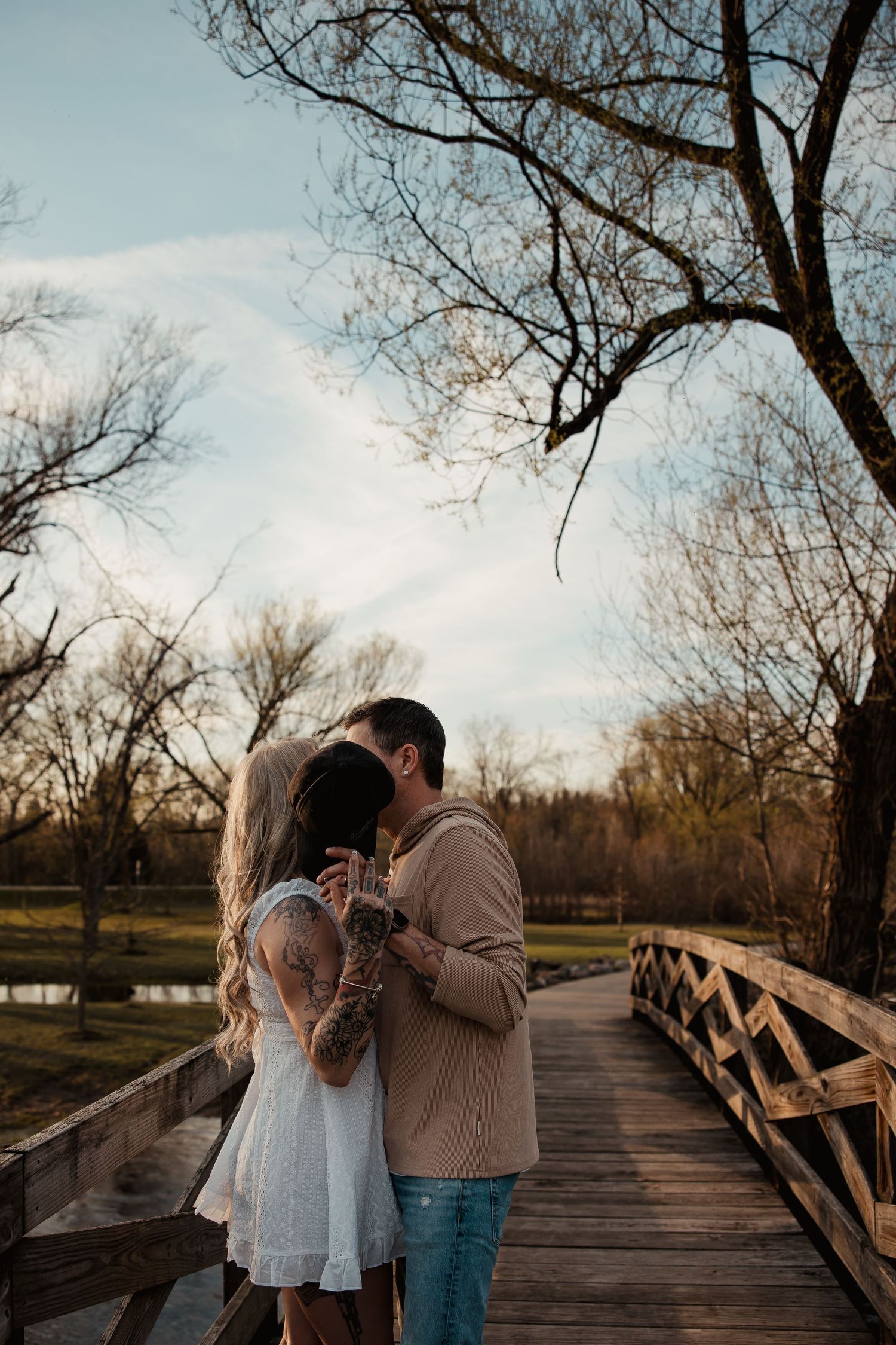 Couple sharing a romantic kiss on a wooden bridge surrounded by bare trees at golden hour.