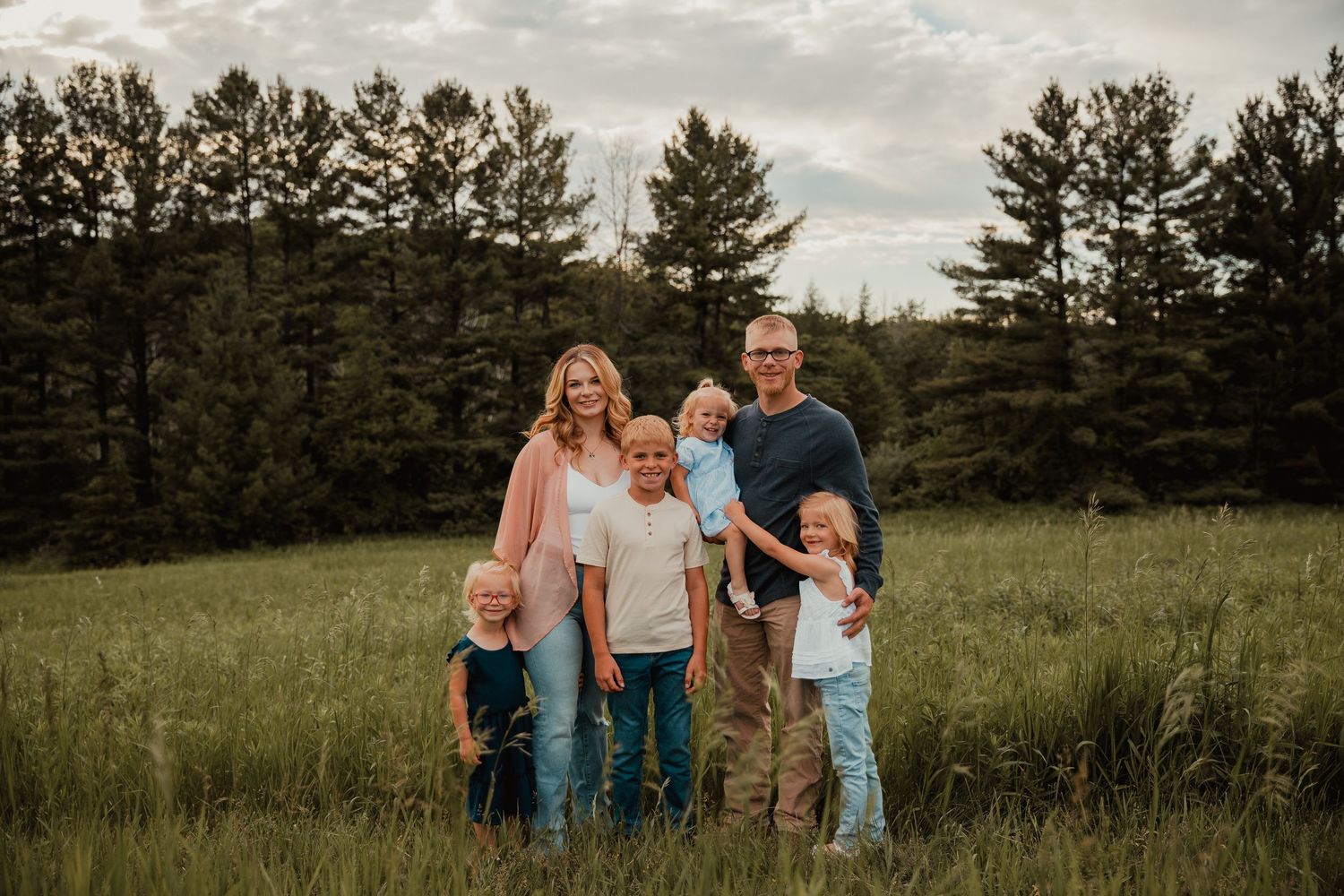 Family of six posing in a lush green field surrounded by tall pine trees during a golden hour outdoor portrait session.