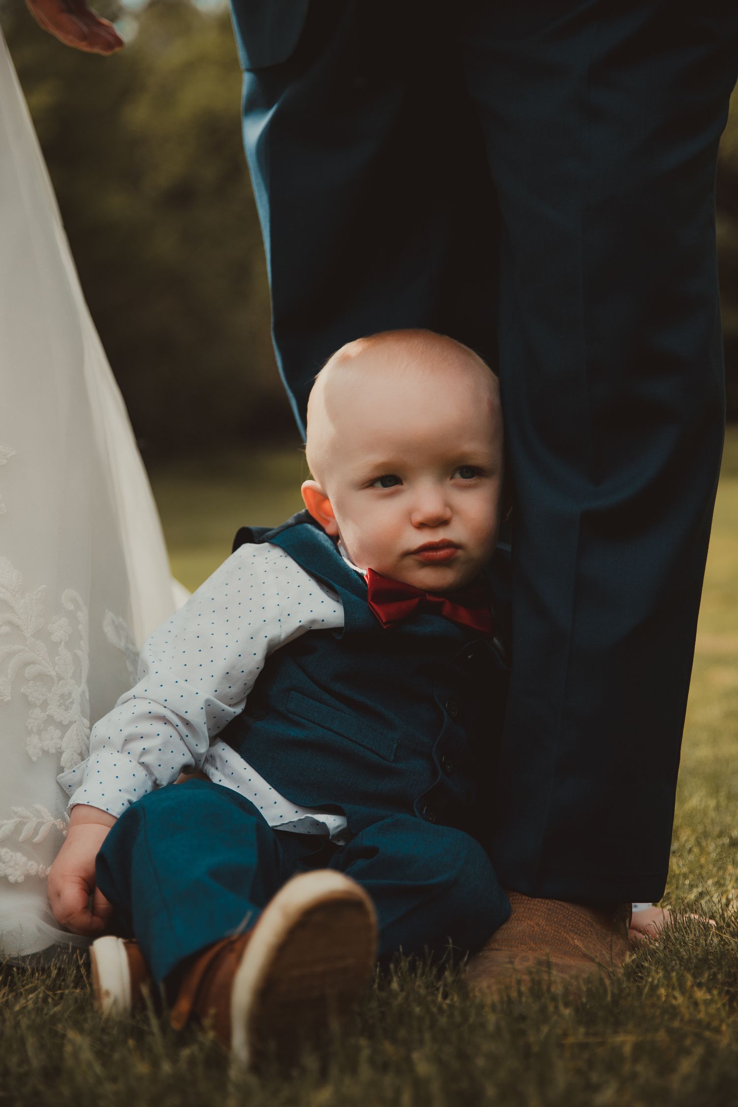 A small child in navy and gray clothing sits beside adult legs on grass. wedding
