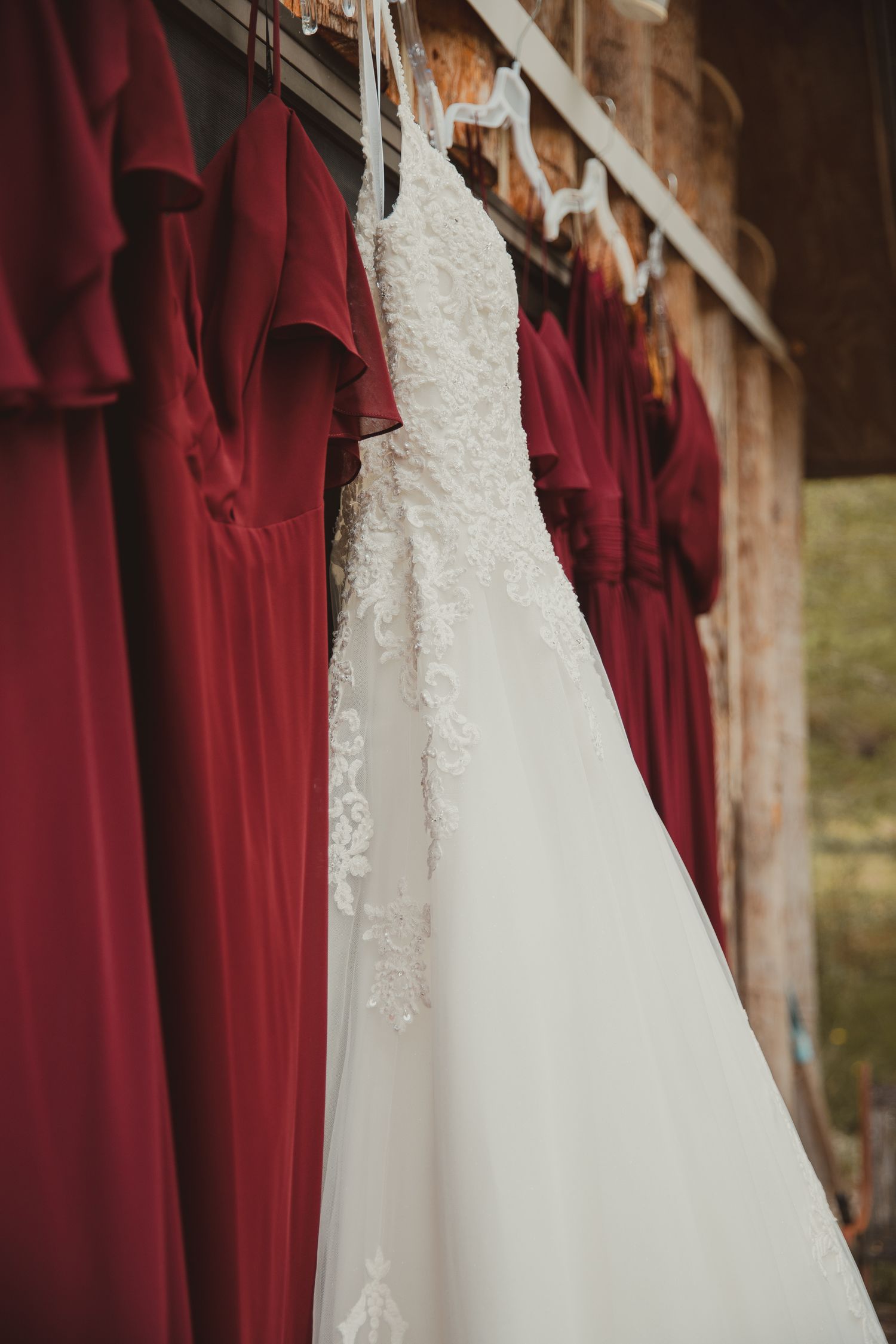 A white lace wedding dress hangs next to burgundy bridesmaid dresses on a rustic wooden wall. wedding