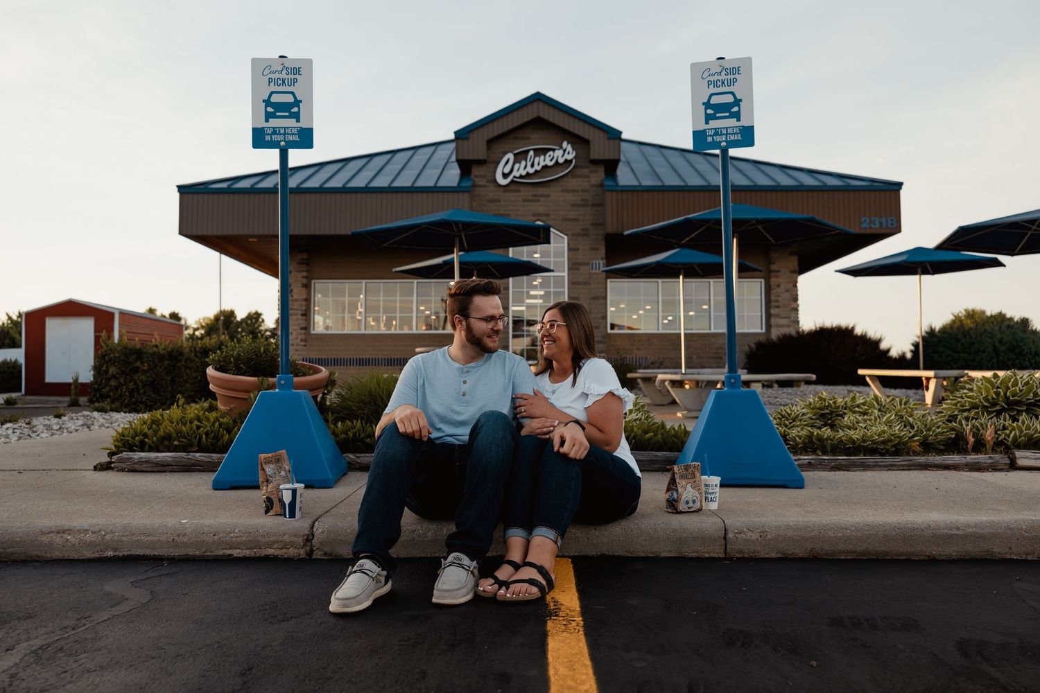 A couple sitting together on a curb in front of a Culvers restaurant smiling.