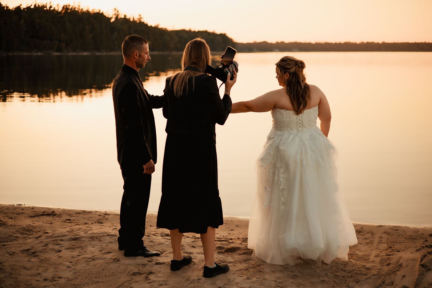 A bride and groom stand on a beach at sunset, with a person officiating their wedding ceremony.
