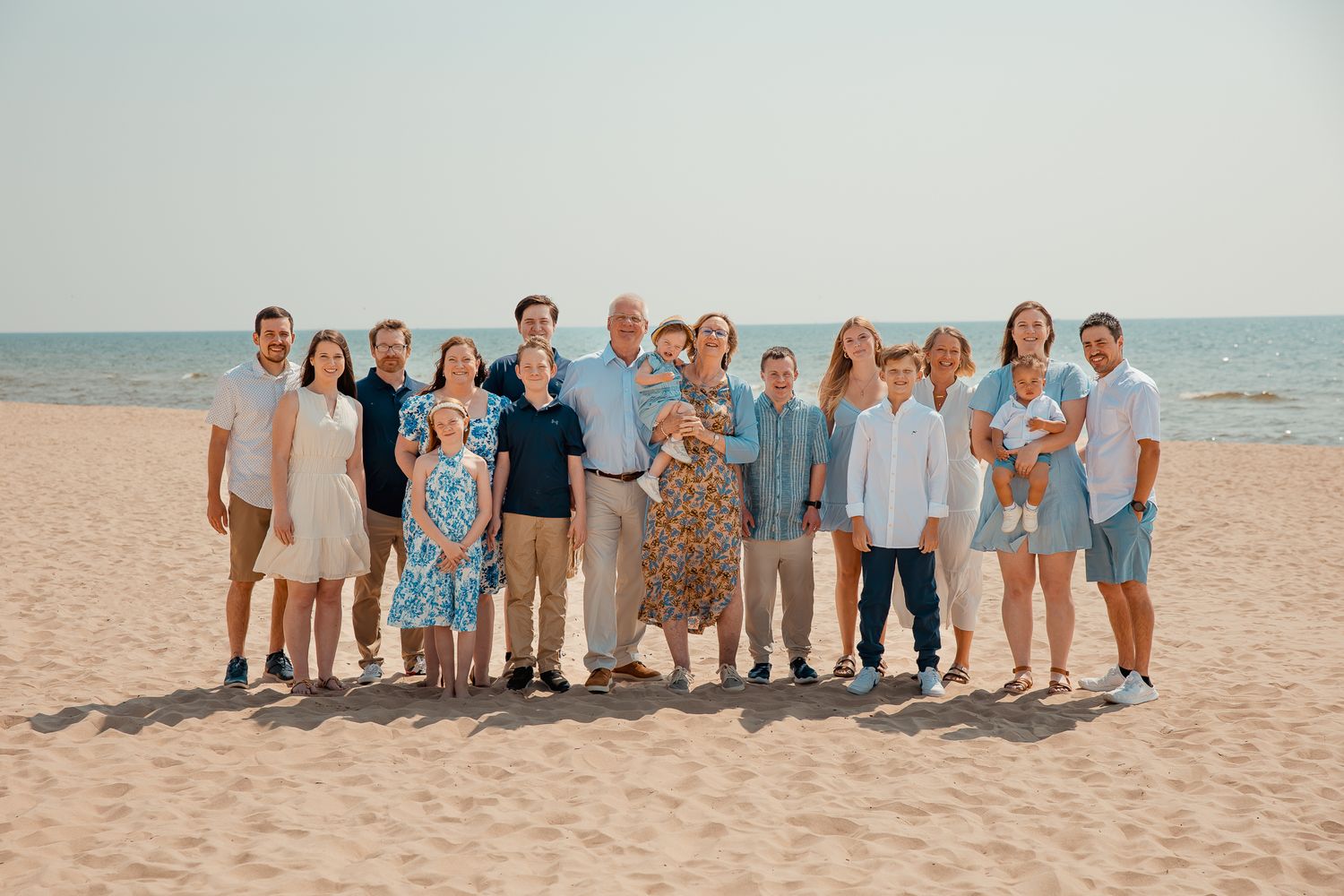 Large family group portrait on a sandy beach with calm ocean waters and clear sky in the background.