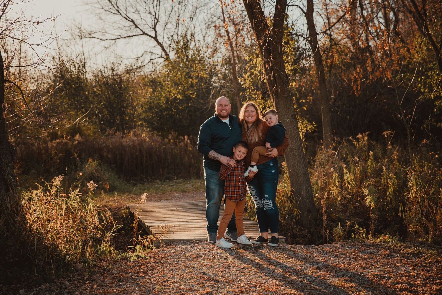 Family of four walking on a leaf-covered path surrounded by autumn trees in golden hour light.