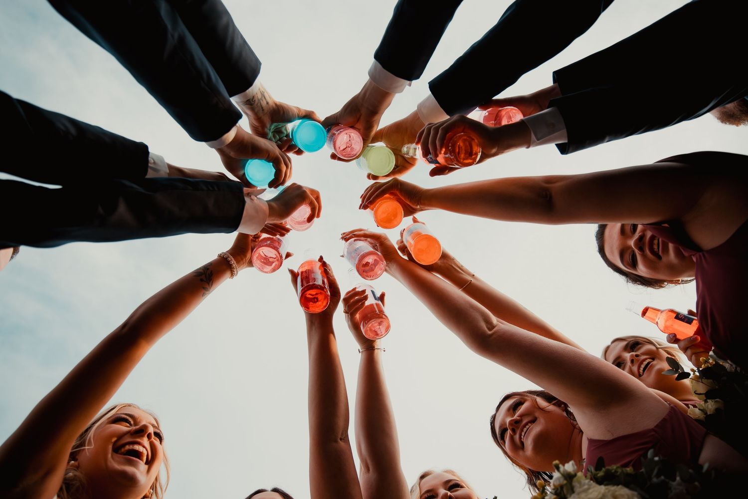 Wedding party toasts with colorful drinks in a circular formation shot from below against blue sky. wedding