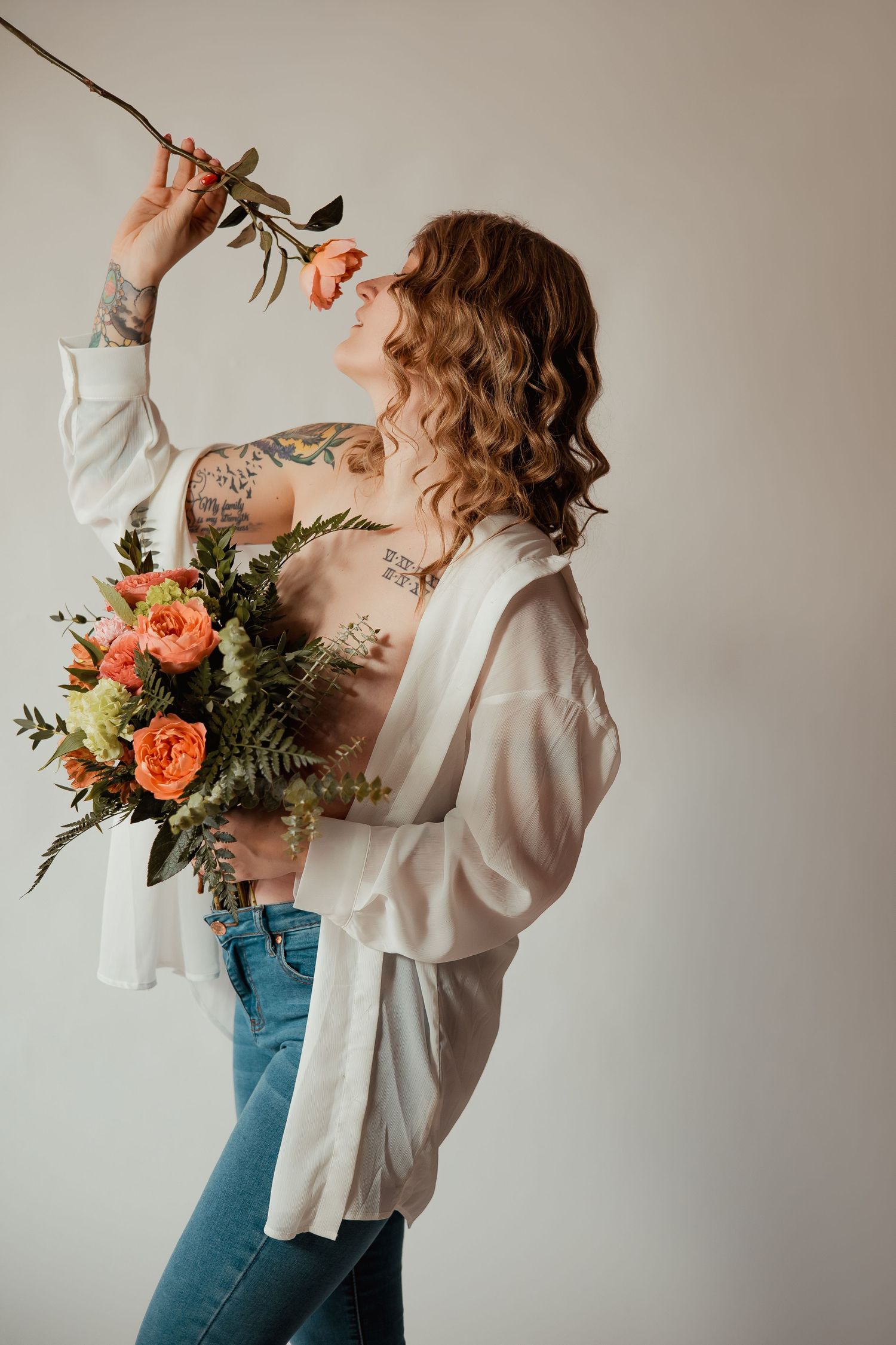 Person in white blouse and jeans holding coral floral bouquet with greenery against neutral wall.