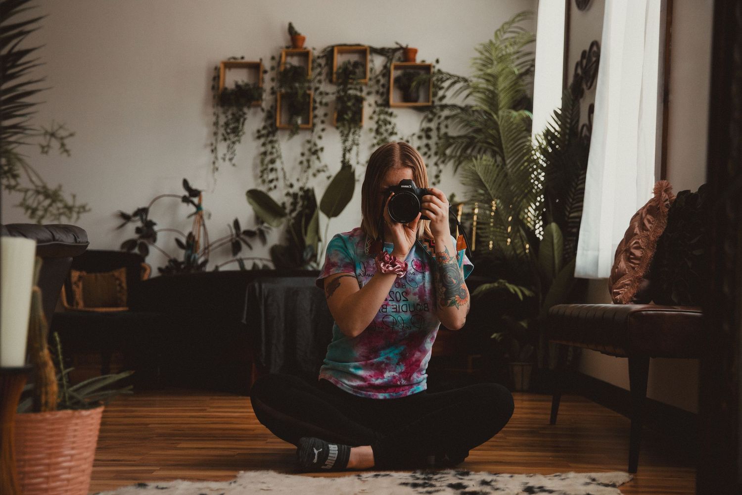 A person sits in a cozy loft area surrounded by plants while working on a laptop near a window.