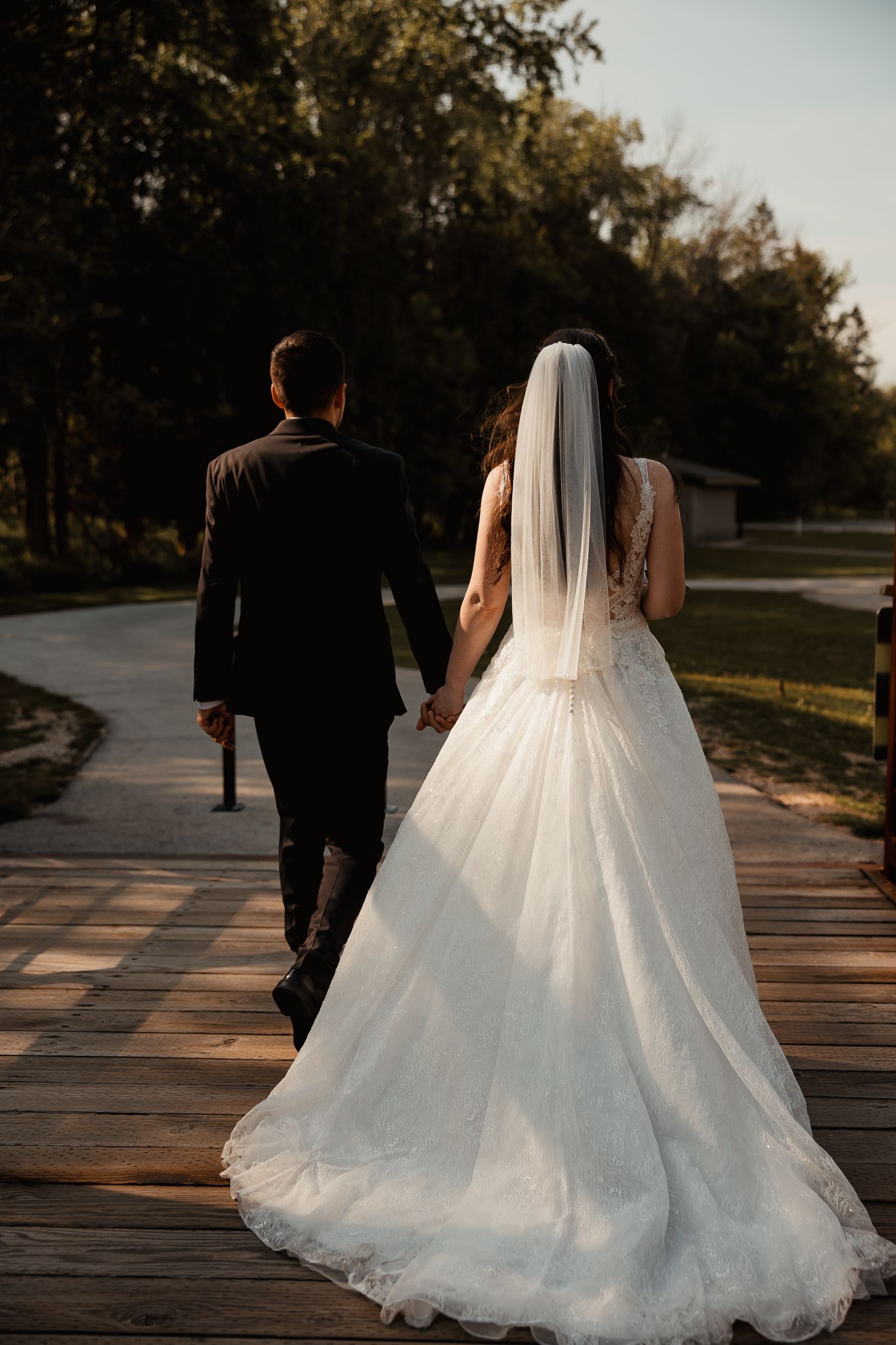 A couple walks down a wooden boardwalk during their wedding, with the bride's white gown train flowing behind them.