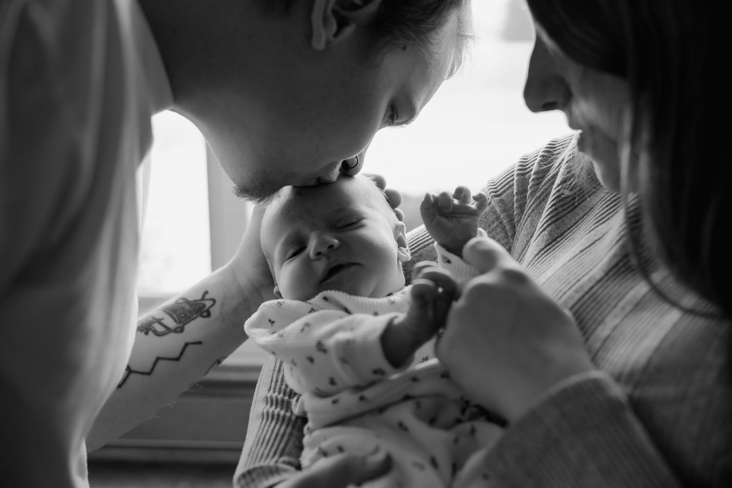 A black and white photo shows a tender moment between a parent and newborn baby in soft window light.