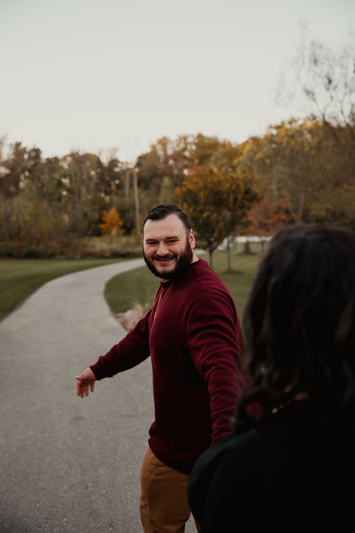 Person in burgundy sweater walking on curved path surrounded by autumn trees at sunset. couples engagement