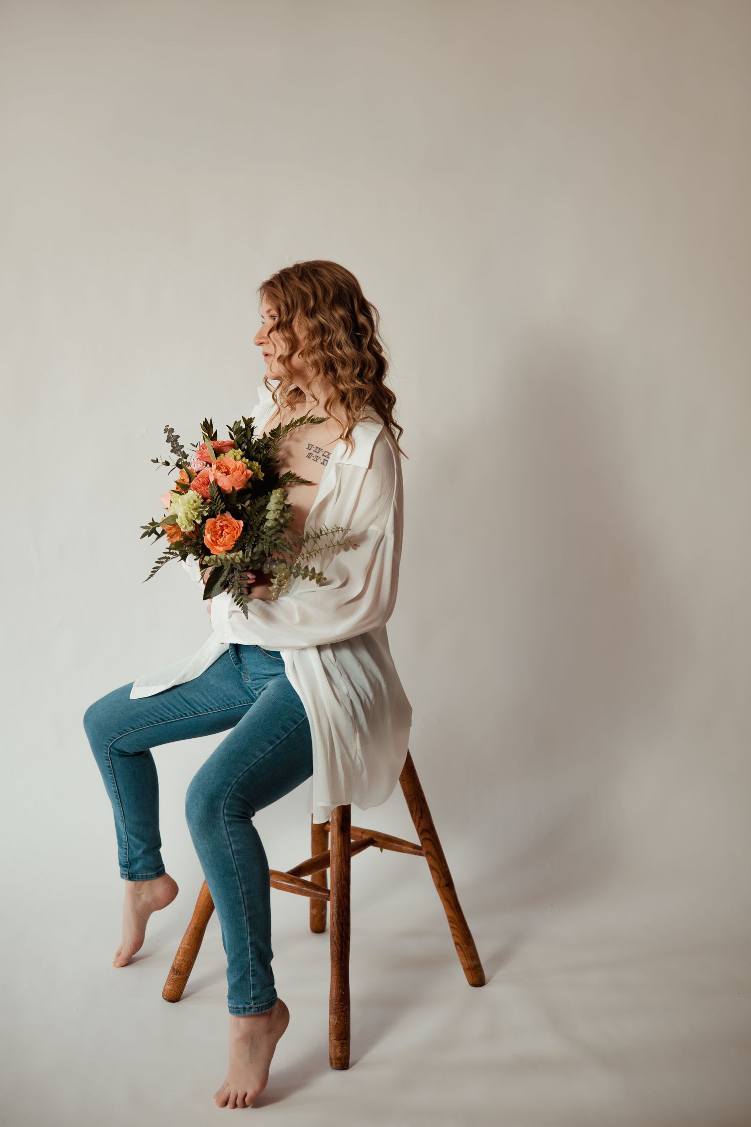 Minimalist studio portrait with wooden stool, white top, and denim styling against beige backdrop.