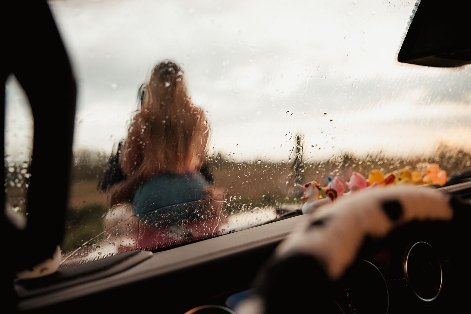 View through a rain-covered car windshield showing a blurry silhouette and stormy sky at sunset.