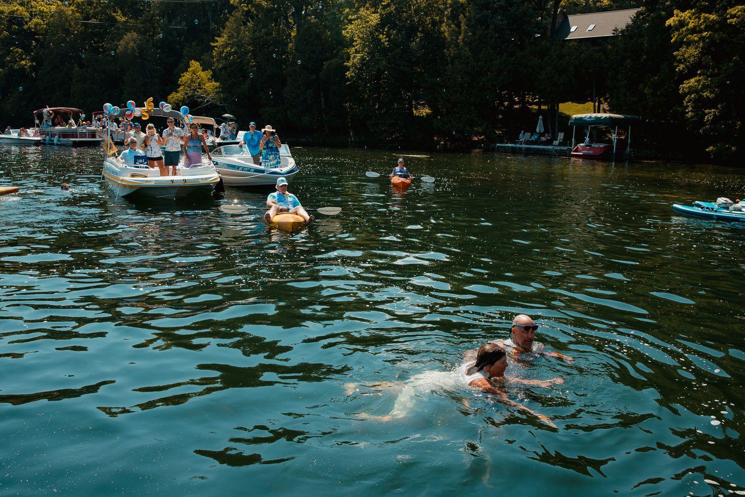 A group of people enjoy a sunny day swimming and paddling in the calm waters of a lake surrounded by trees.