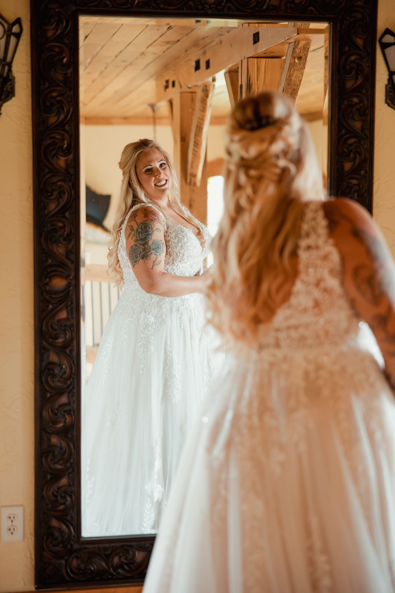 A bride in a white lace wedding dress looks at her reflection in an ornate framed mirror.
