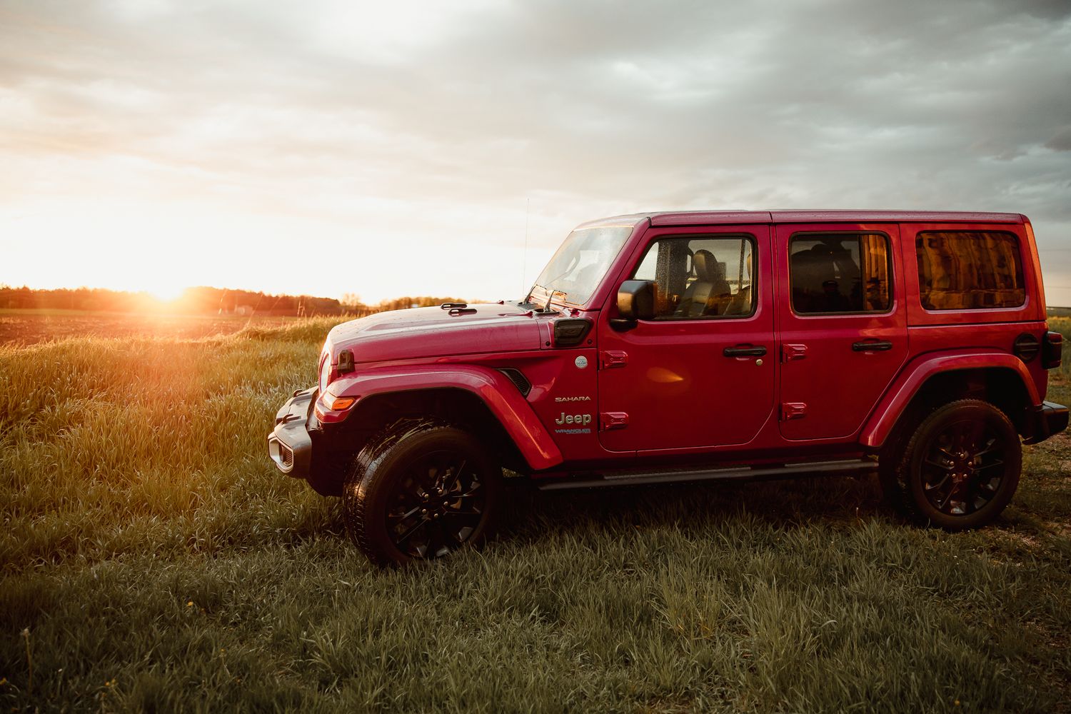 Red Jeep Wrangler parked in a grassy field at sunset with dramatic cloudy sky in the background.