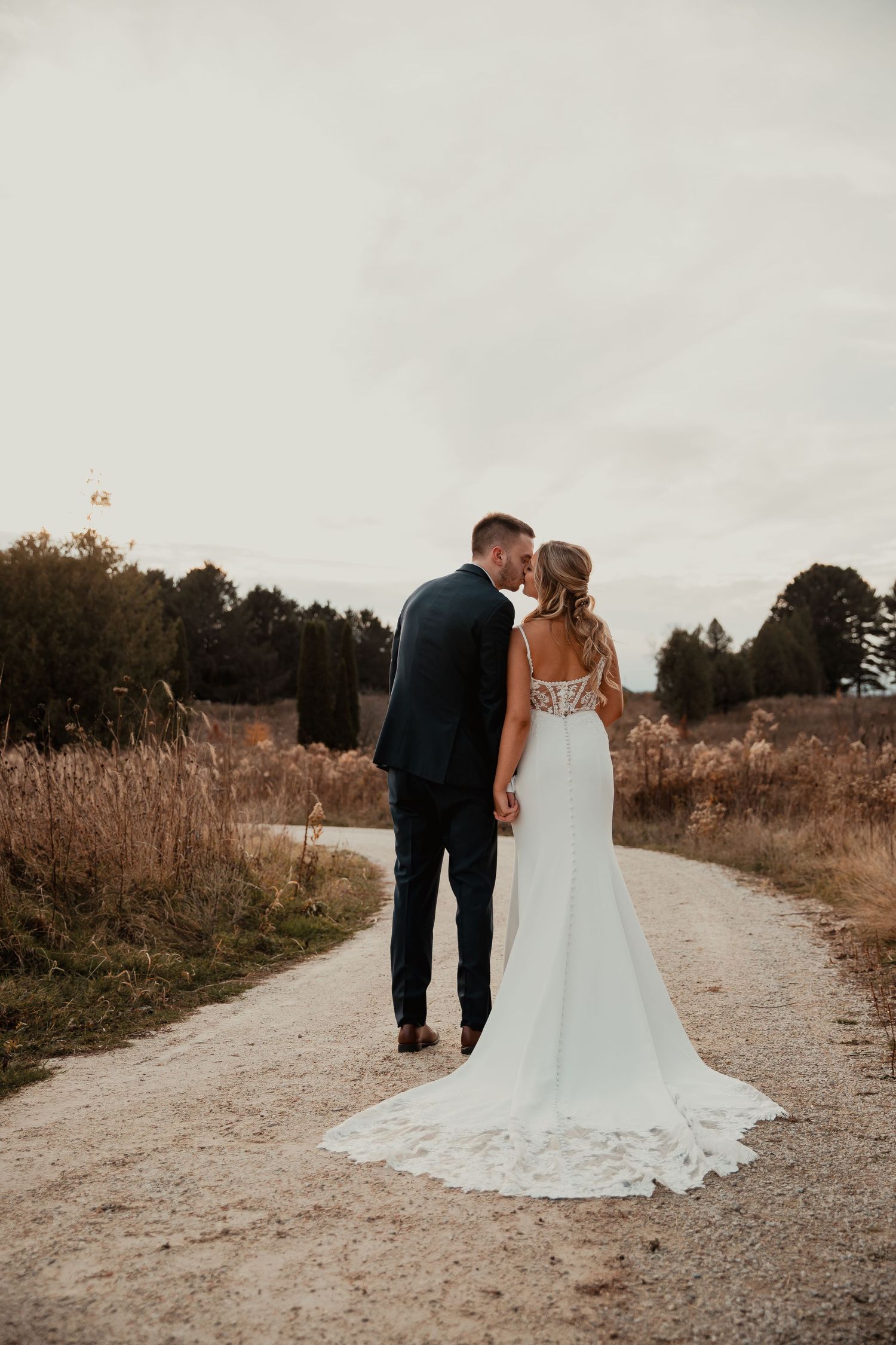 Couple in wedding attire walking down dirt path with long white train trailing behind. wedding