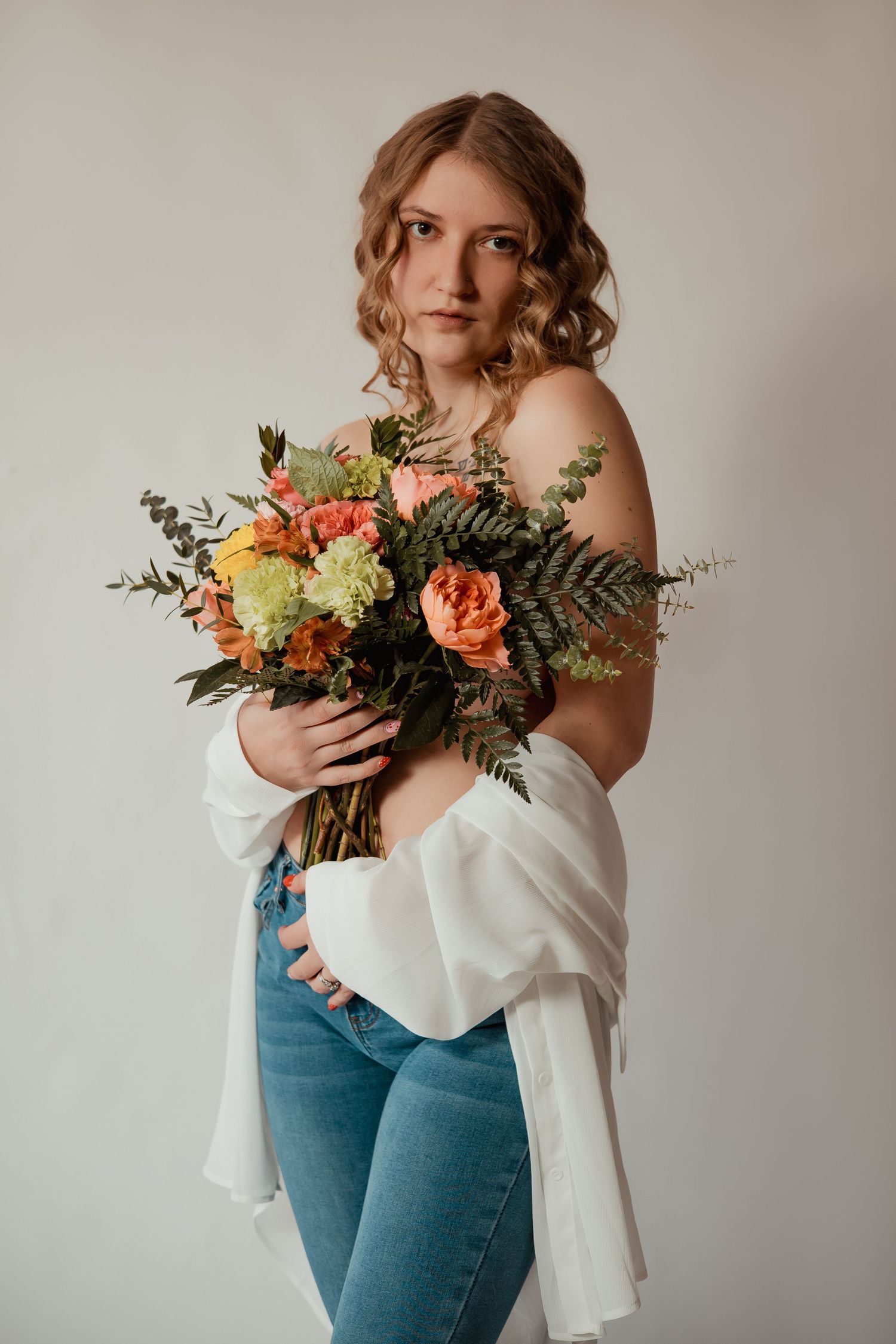 A floral arrangement with peach roses and greenery is held against a white blouse and blue jeans in a minimalist setting.