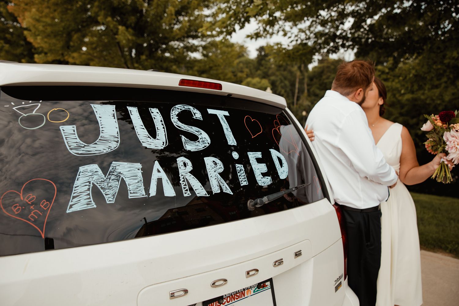 A car window with Just Married written in chalk text while a couple embraces in the background.