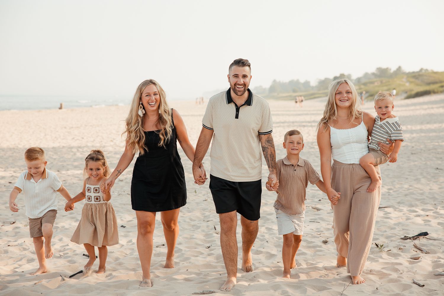 Family of seven walking on a sunny beach, parents holding hands with children, smiling and barefoot on sand.