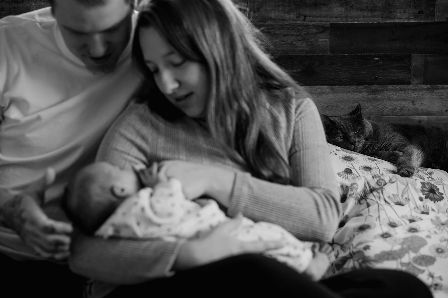 A tender black and white photo shows a couple gazing lovingly at their newborn baby while resting on a bed.