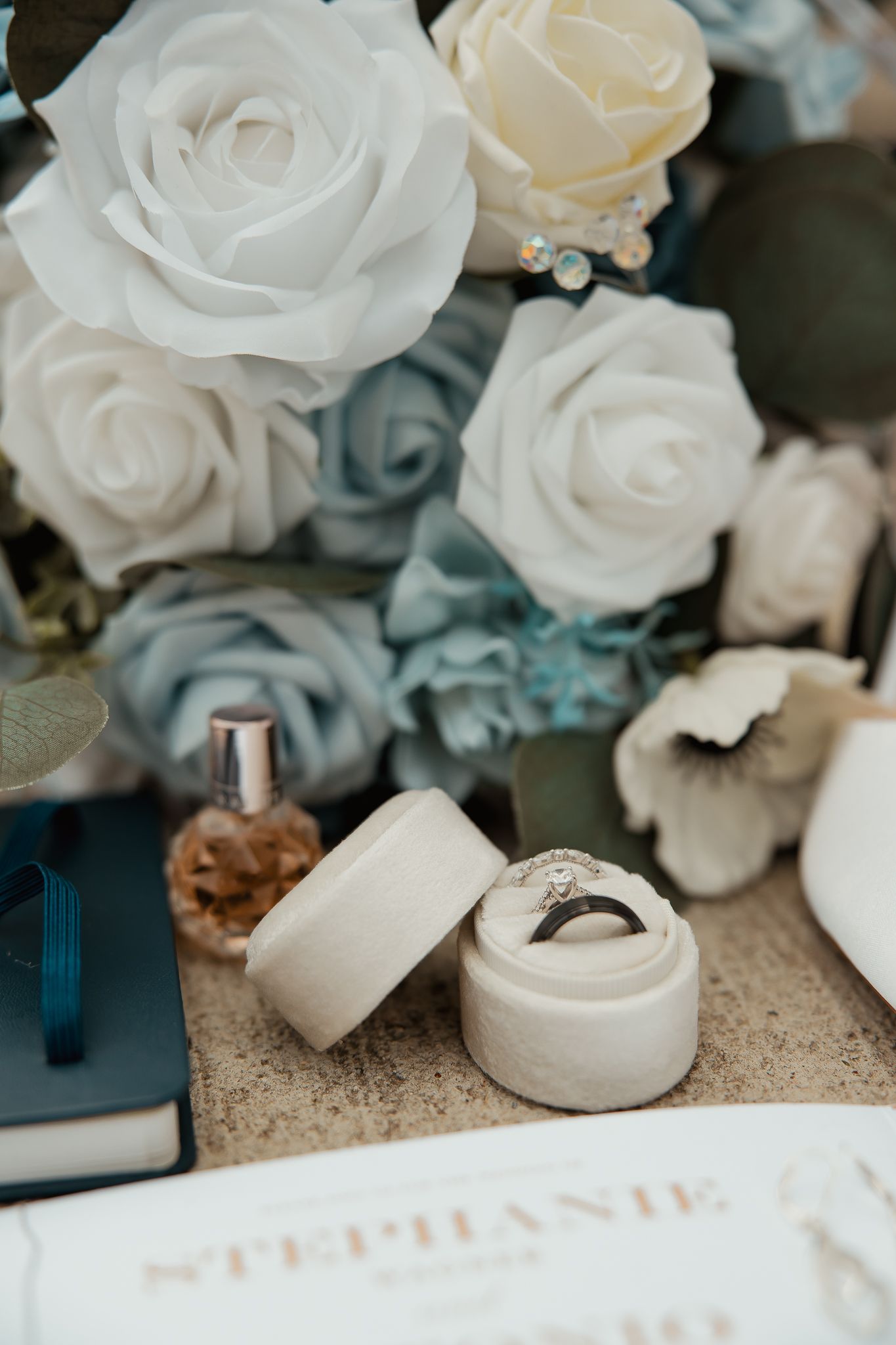 White rose bouquet and ring boxes arranged on beige surface with blue accents for wedding photography.