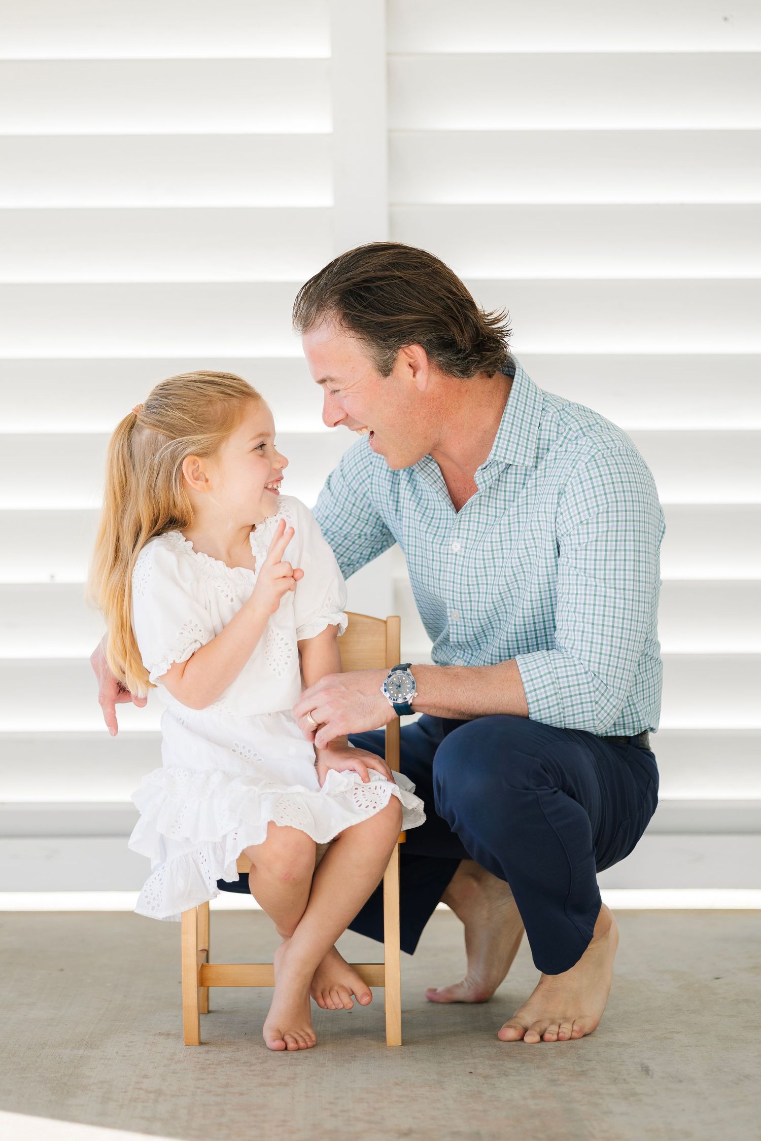 A father crouches down to chat and laugh with his young blonde daughter seated on a small wooden chair.