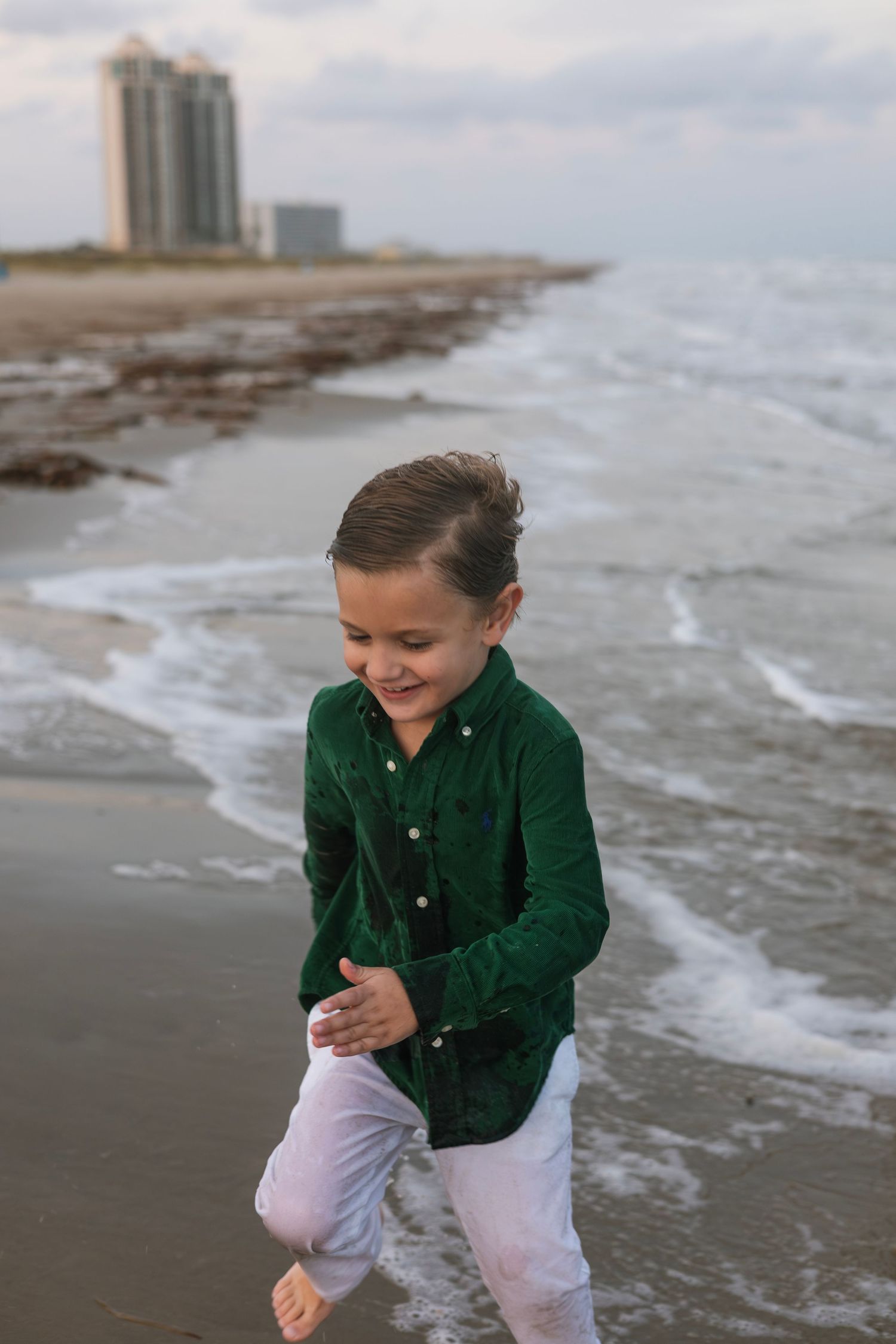 Young child in green sweater plays joyfully along a sandy beach shoreline during a cloudy day.