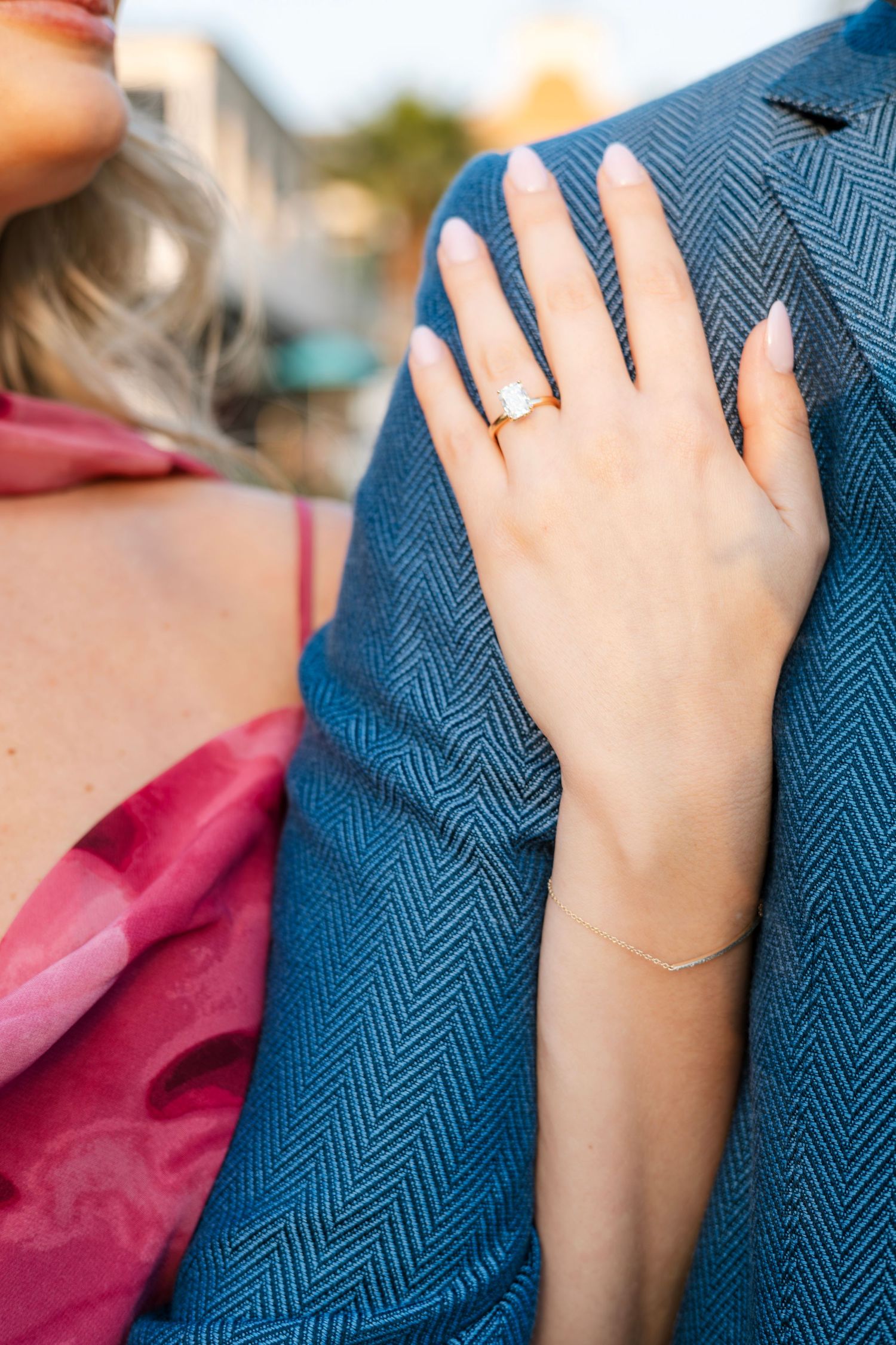Close-up of a woman's hand with a gold diamond engagement ring resting on a man's blue herringbone suit jacket.