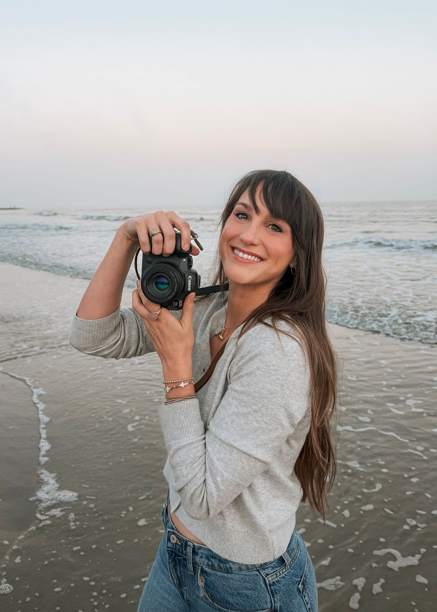 Smiling woman holding a camera at the beach during golden hour, wearing a gray top and jeans.