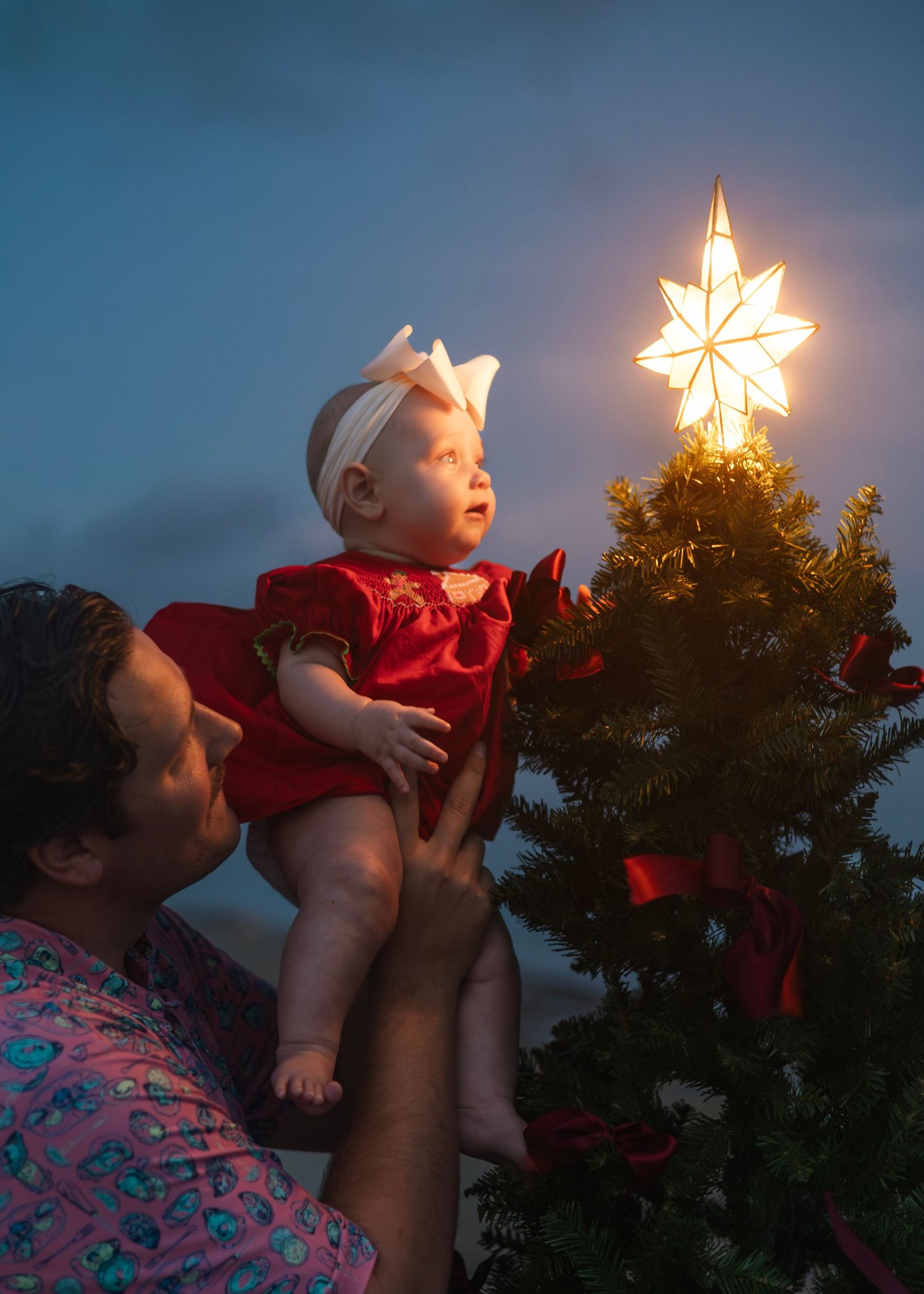 A parent lifts a baby wearing red clothing to reach a glowing star decoration atop a Christmas tree at dusk.