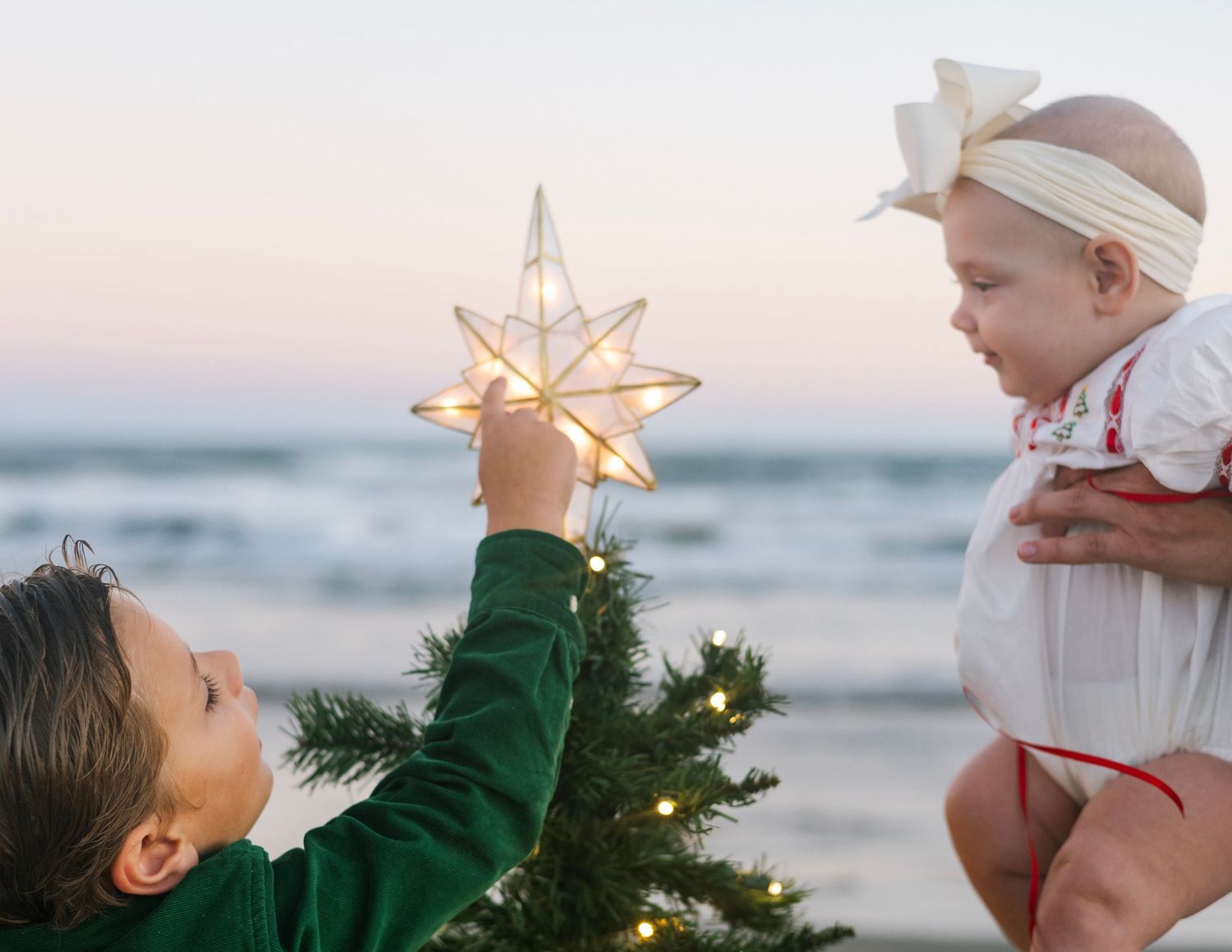 A family places a glowing star ornament on top of a Christmas tree at the beach during sunset.