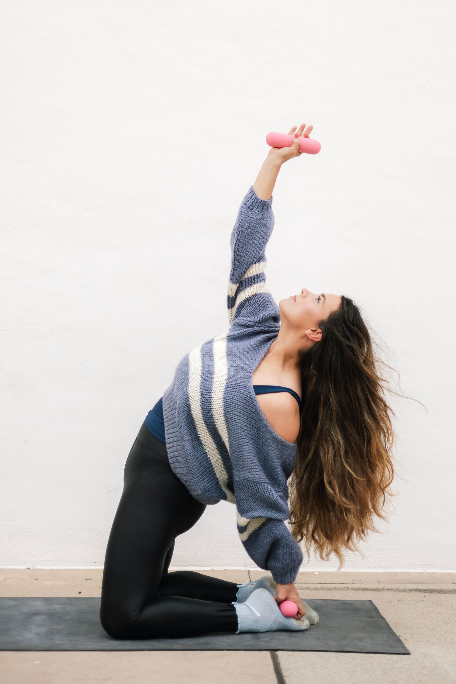 Woman in gray sweater and black leggings exercises on yoga mat, lifting pink dumbbell overhead in a kneeling side stretch.