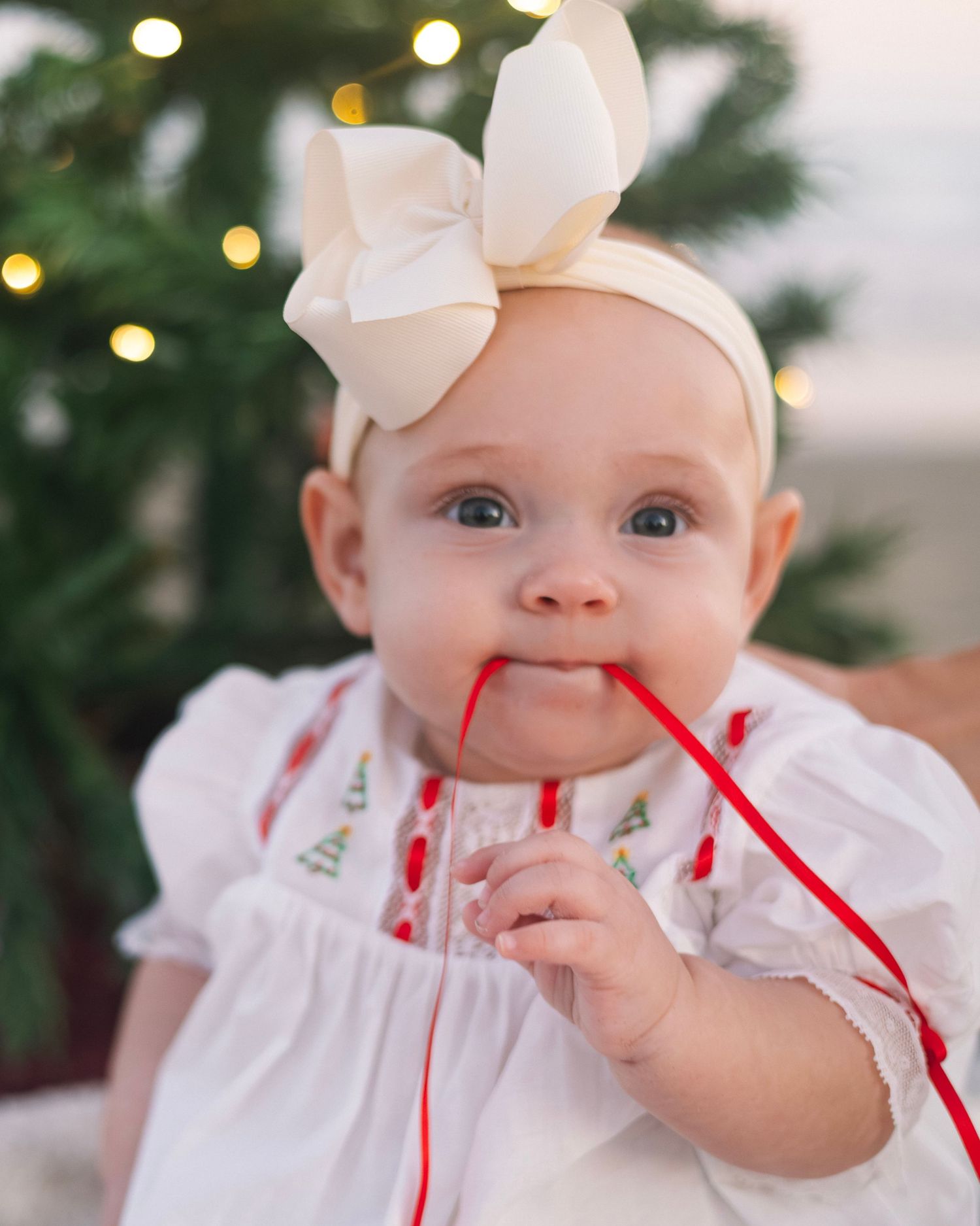 Baby in white dress with red accents and bow headband looking at holiday lights.