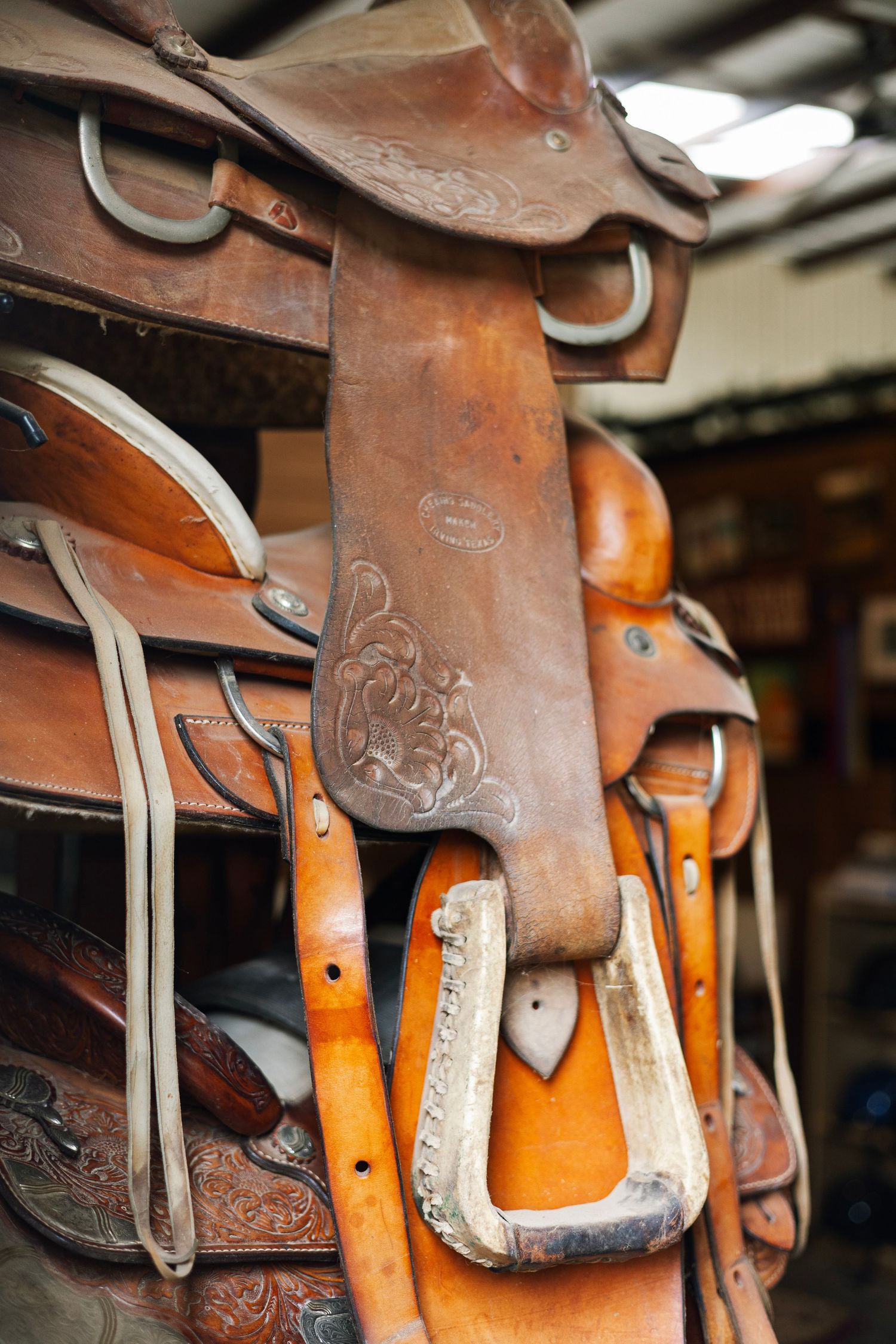 Stack of ornate brown leather Western saddles with stirrups displayed on a rack in a tack shop.