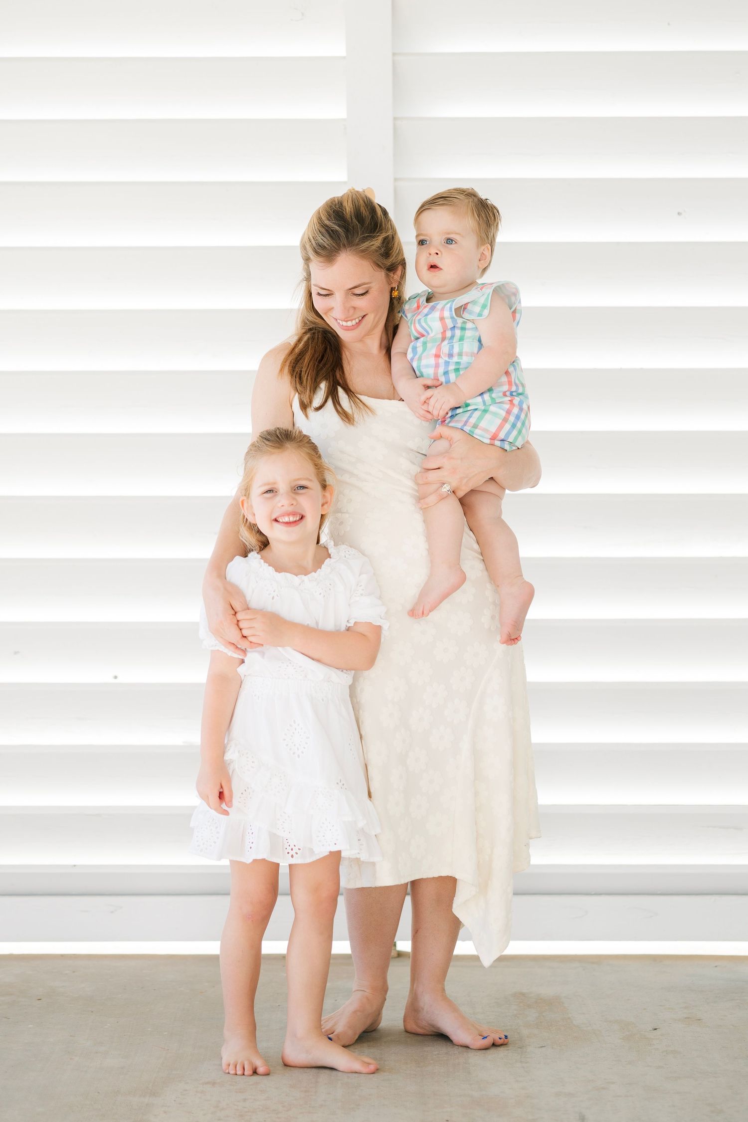 A smiling mother in a white dress holds her baby while her young daughter in a white dress hugs her leg indoors.