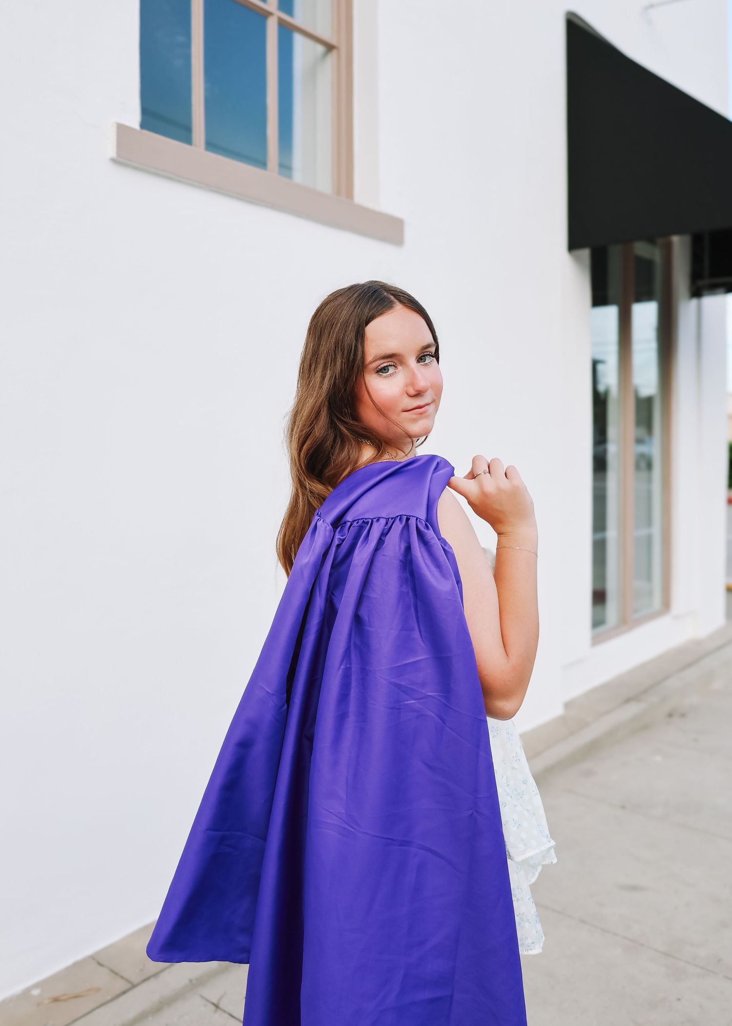 A person in a purple dress poses against a white building wall.