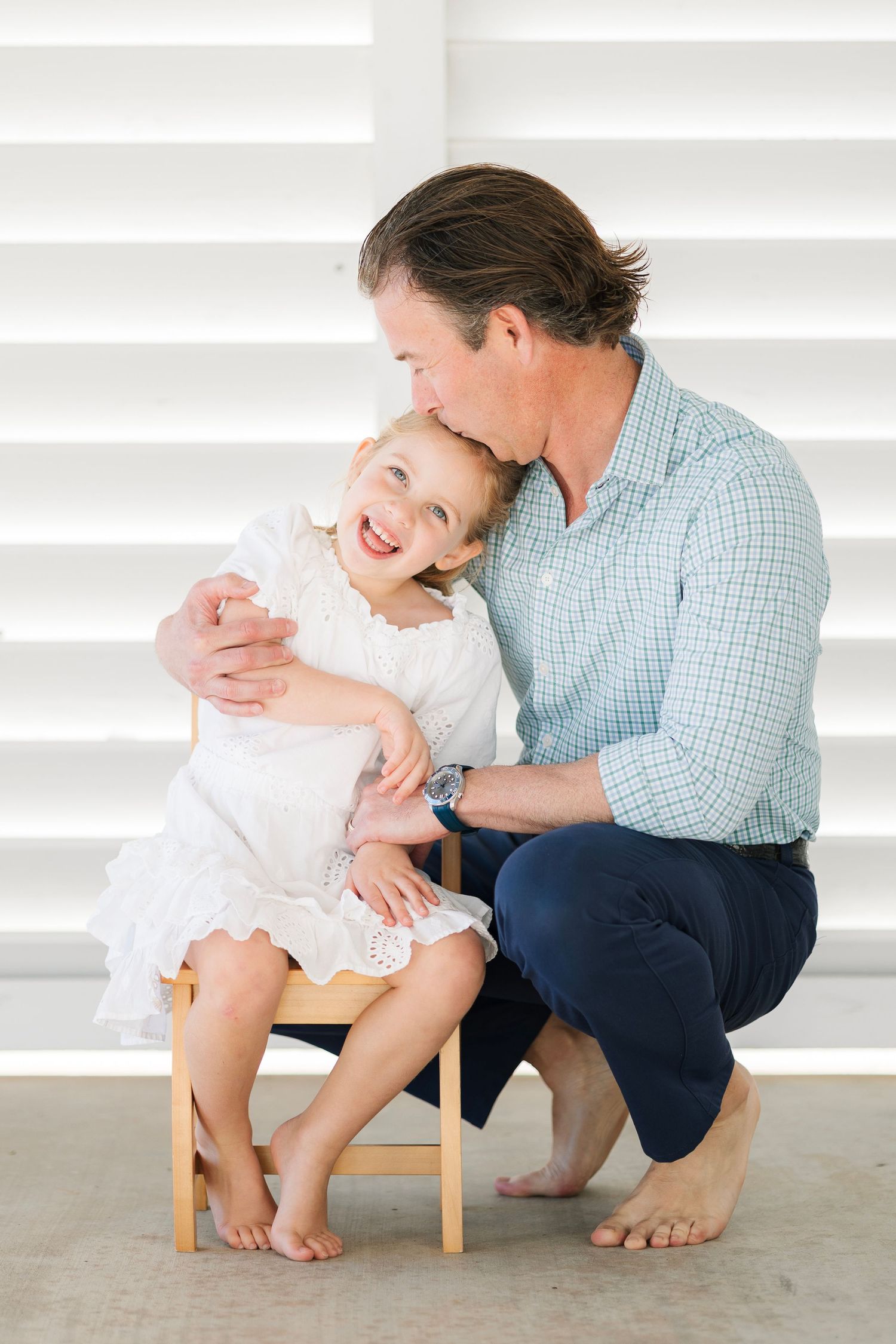 A father kisses his laughing toddler daughter in a white dress while she sits on a small wooden chair.