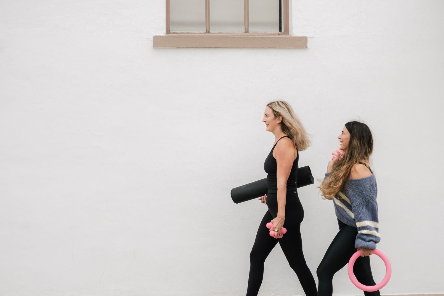 Two women in activewear walking with yoga mats, heading to a fitness class outdoors.