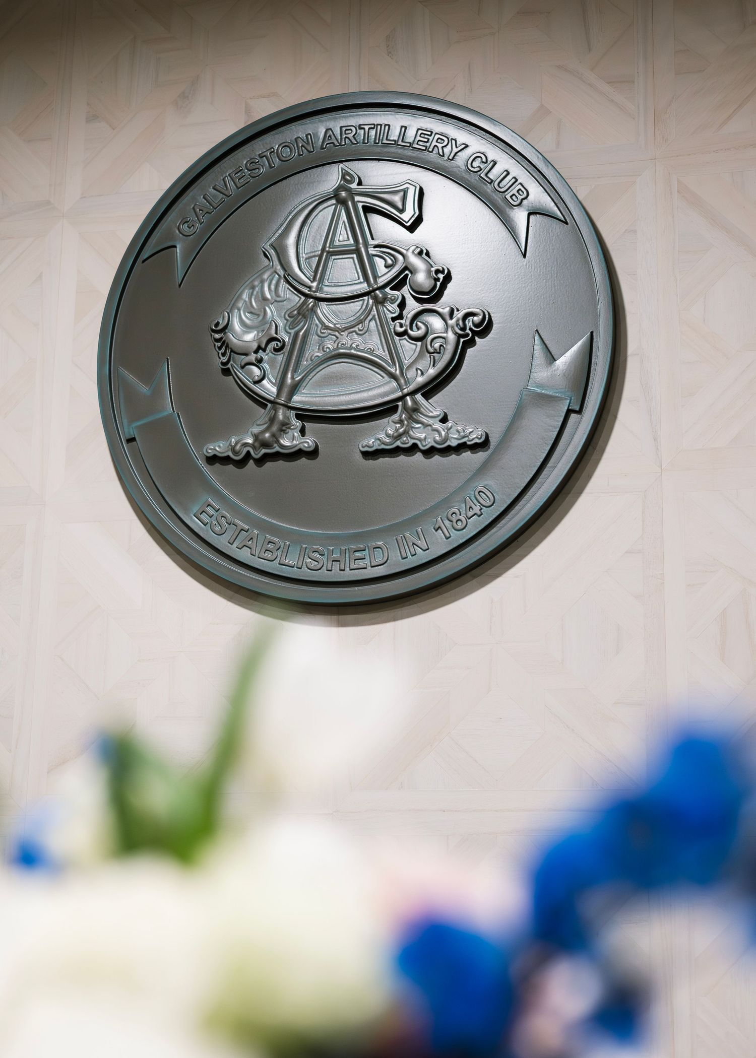 Silver metallic medallion with decorative embossed design displayed against a textured beige background.