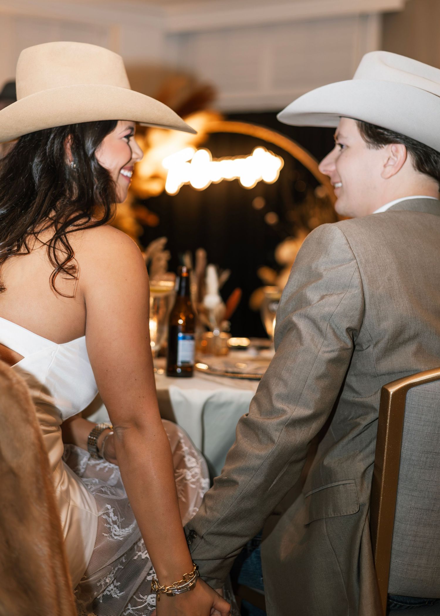 Two newlyweds in white cowboy hats hold hands at their western-themed wedding reception dinner table.