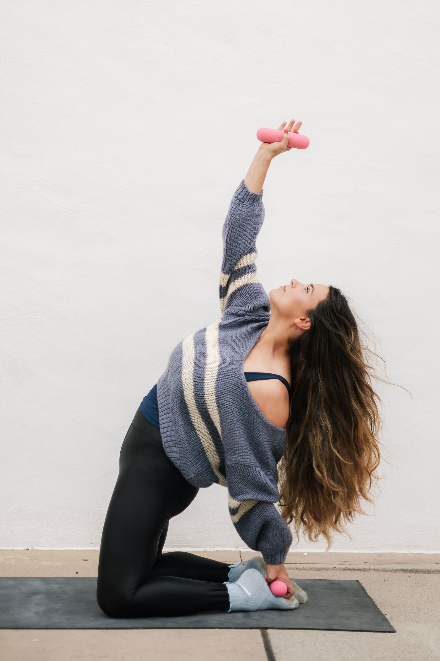 Woman in gray sweater and black leggings kneeling on yoga mat, lifting pink dumbbell overhead in a side stretch exercise.